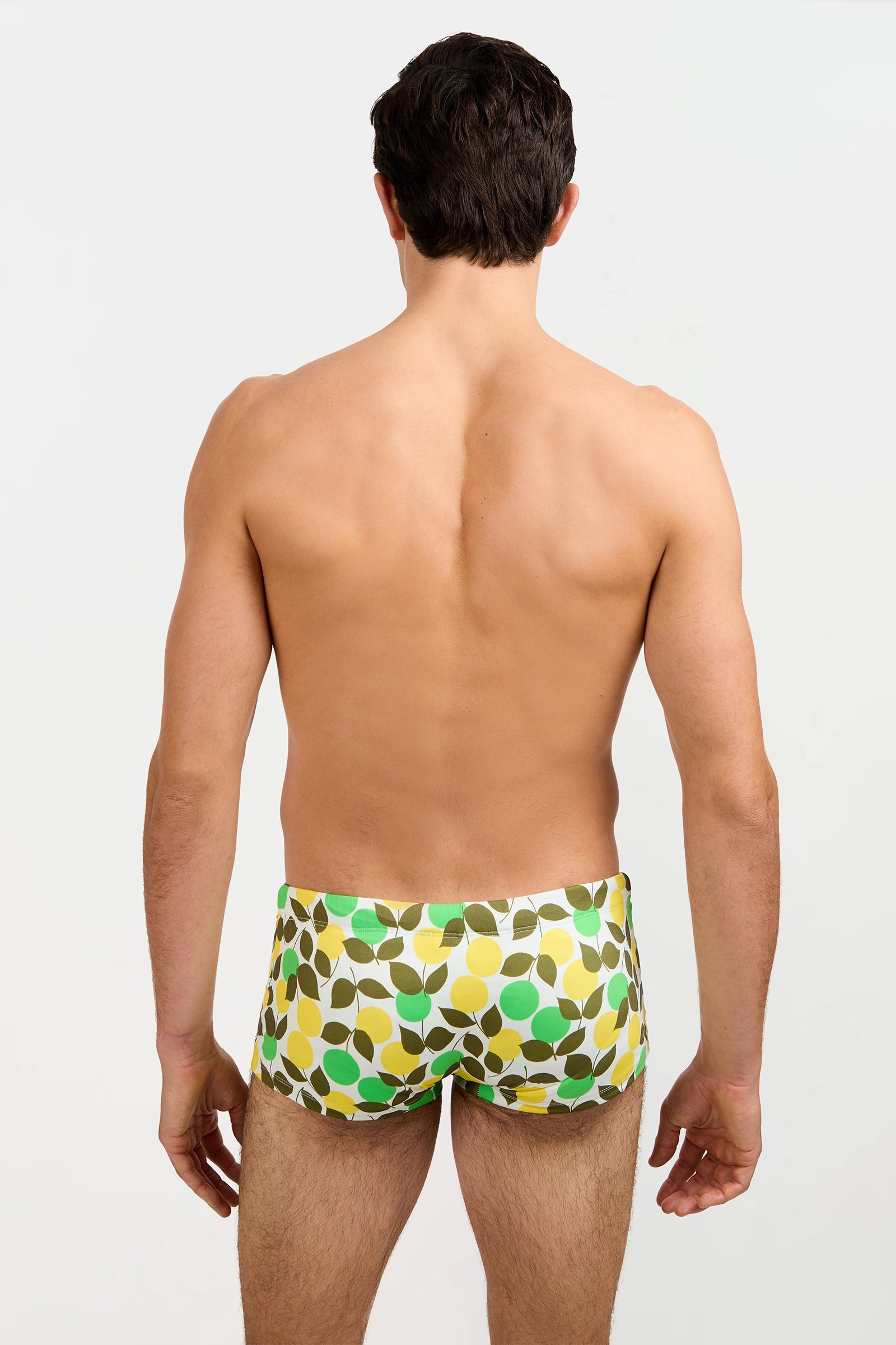 A man faces away from the camera, wearing the Sorrento Swim Trunk - Limone with a green, yellow, and white leaf pattern. His bare back is visible against a plain light background.