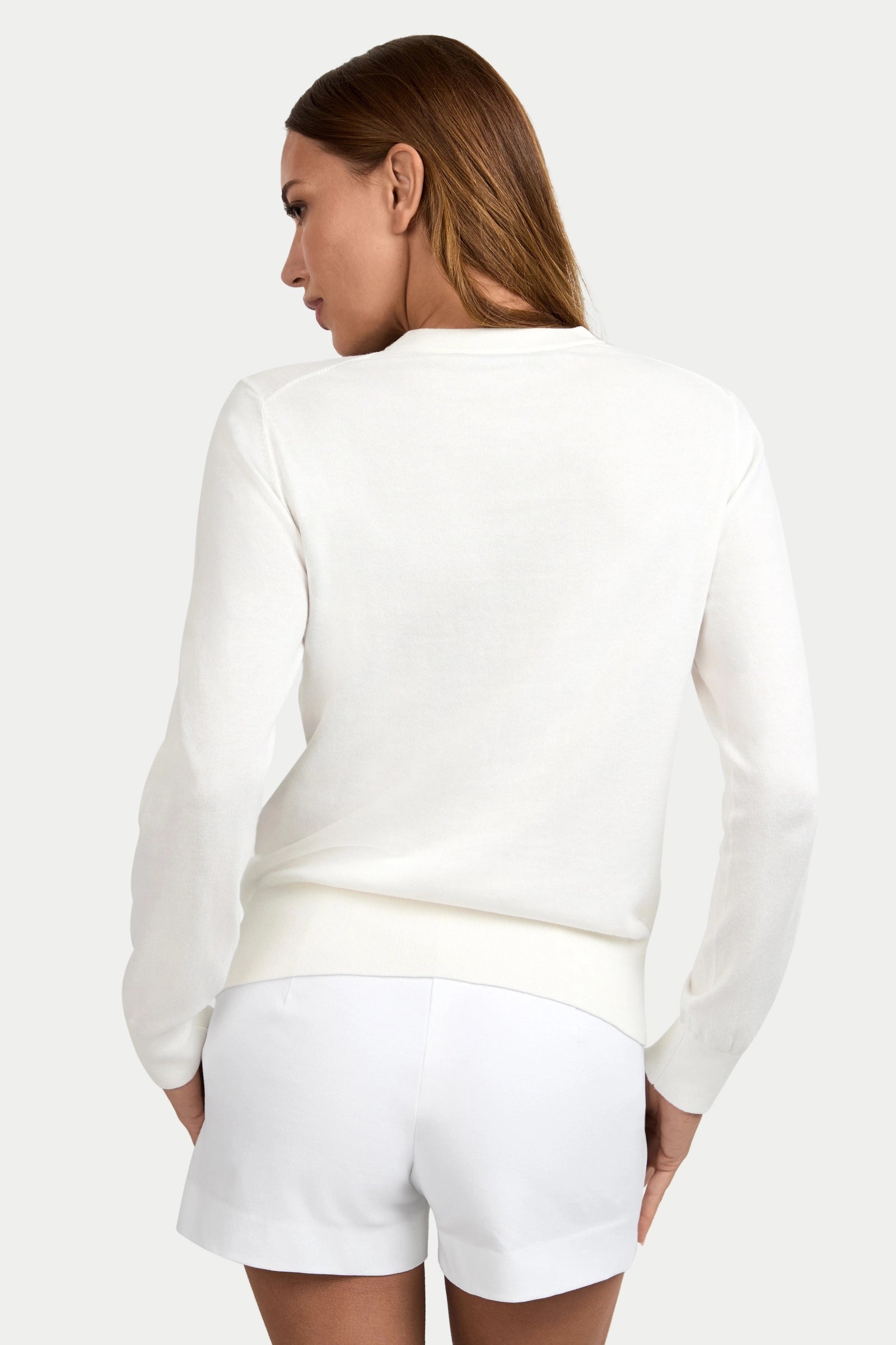 A woman with long brown hair is seen from behind, wearing the Old Fashioned Sweater in white with matching shorts, standing against a plain light background.