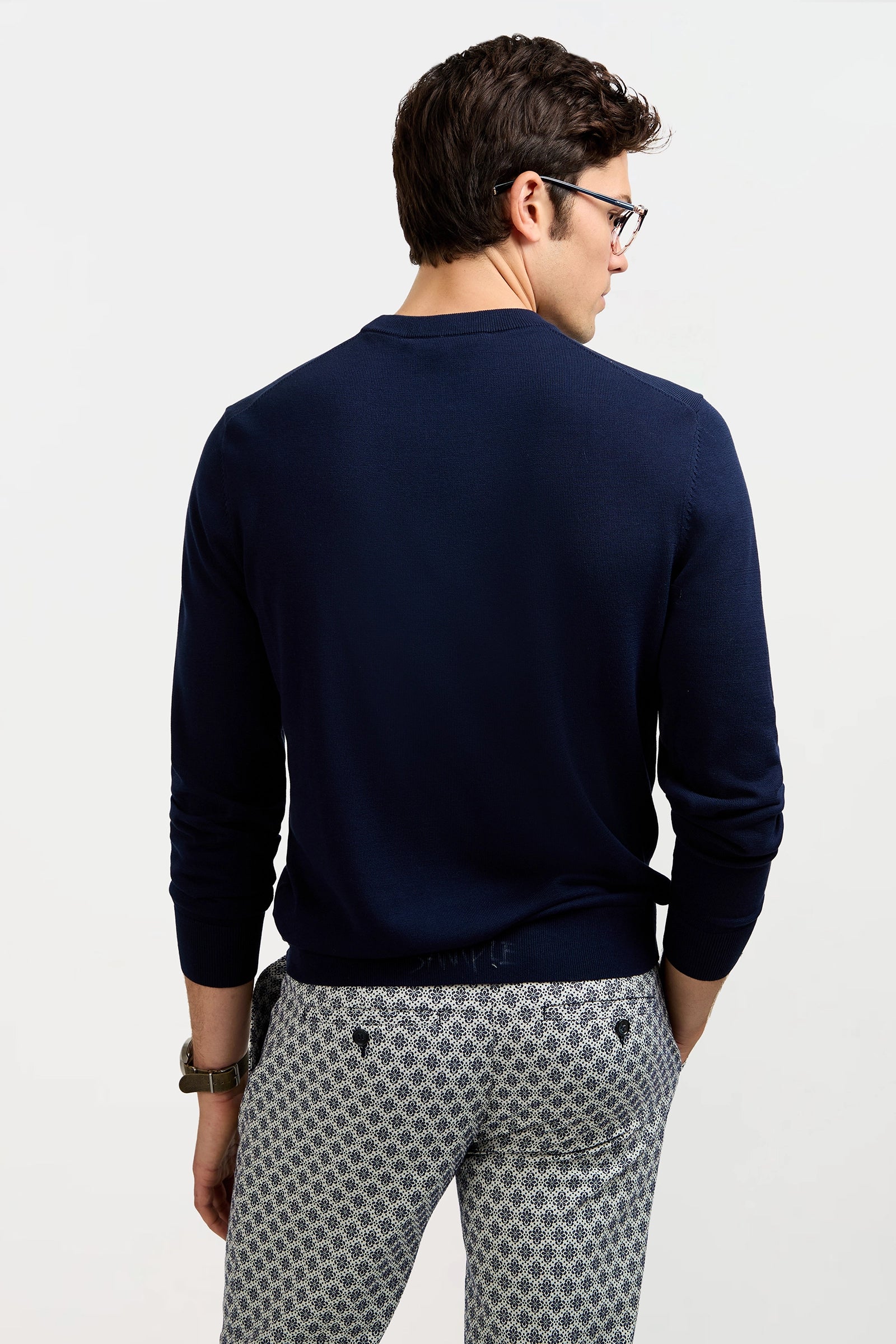 A man wearing the Meyer Long-Sleeve Crewneck Sweater in navy and patterned gray pants stands with his back to the camera. He has short brown hair, glasses, and one hand casually placed in his pocket.