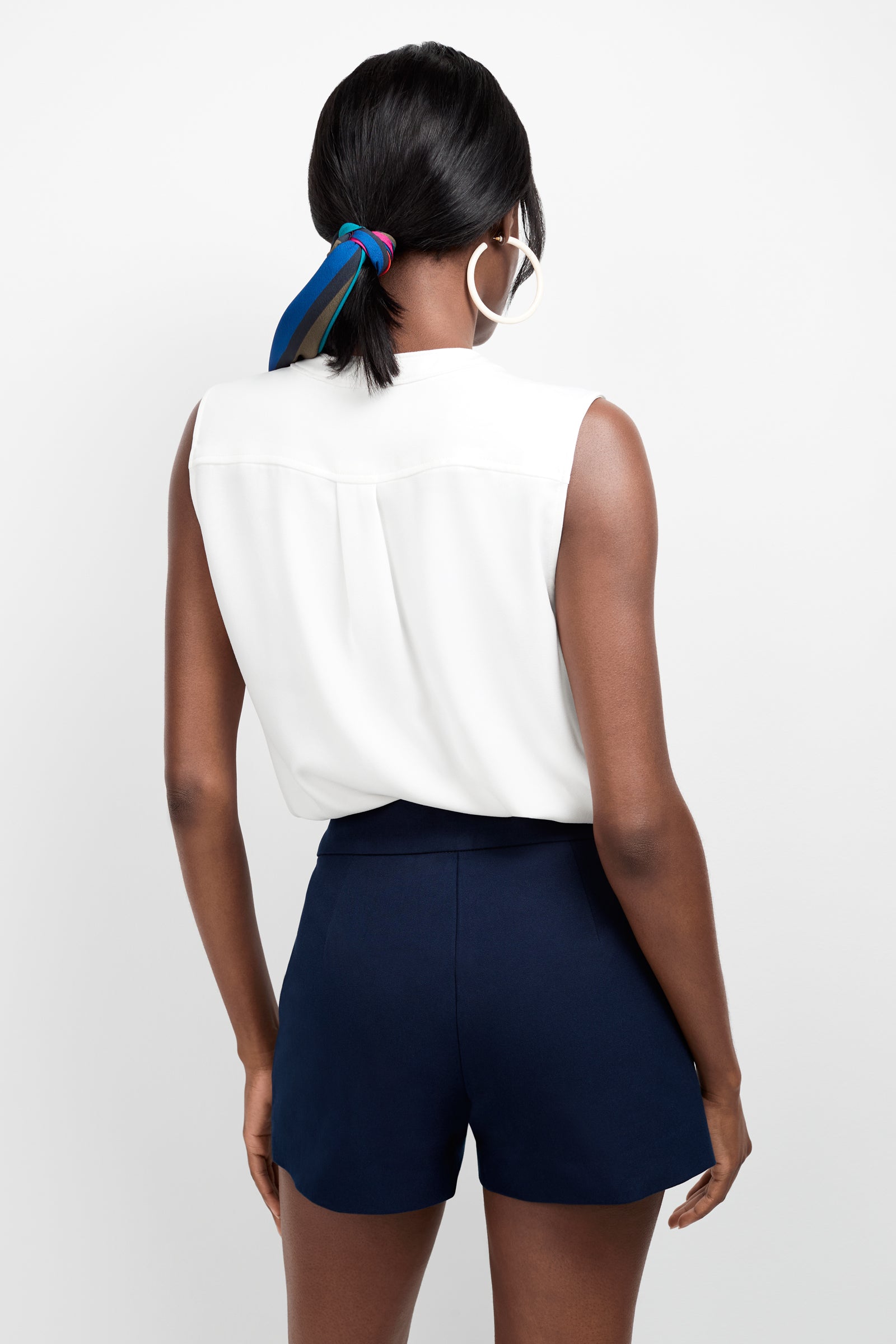 A woman facing away from the camera wears the Sleeveless Kaiko Top, navy high-waisted shorts, large hoop earrings, and a blue striped scarf in her hair against a plain white background.
