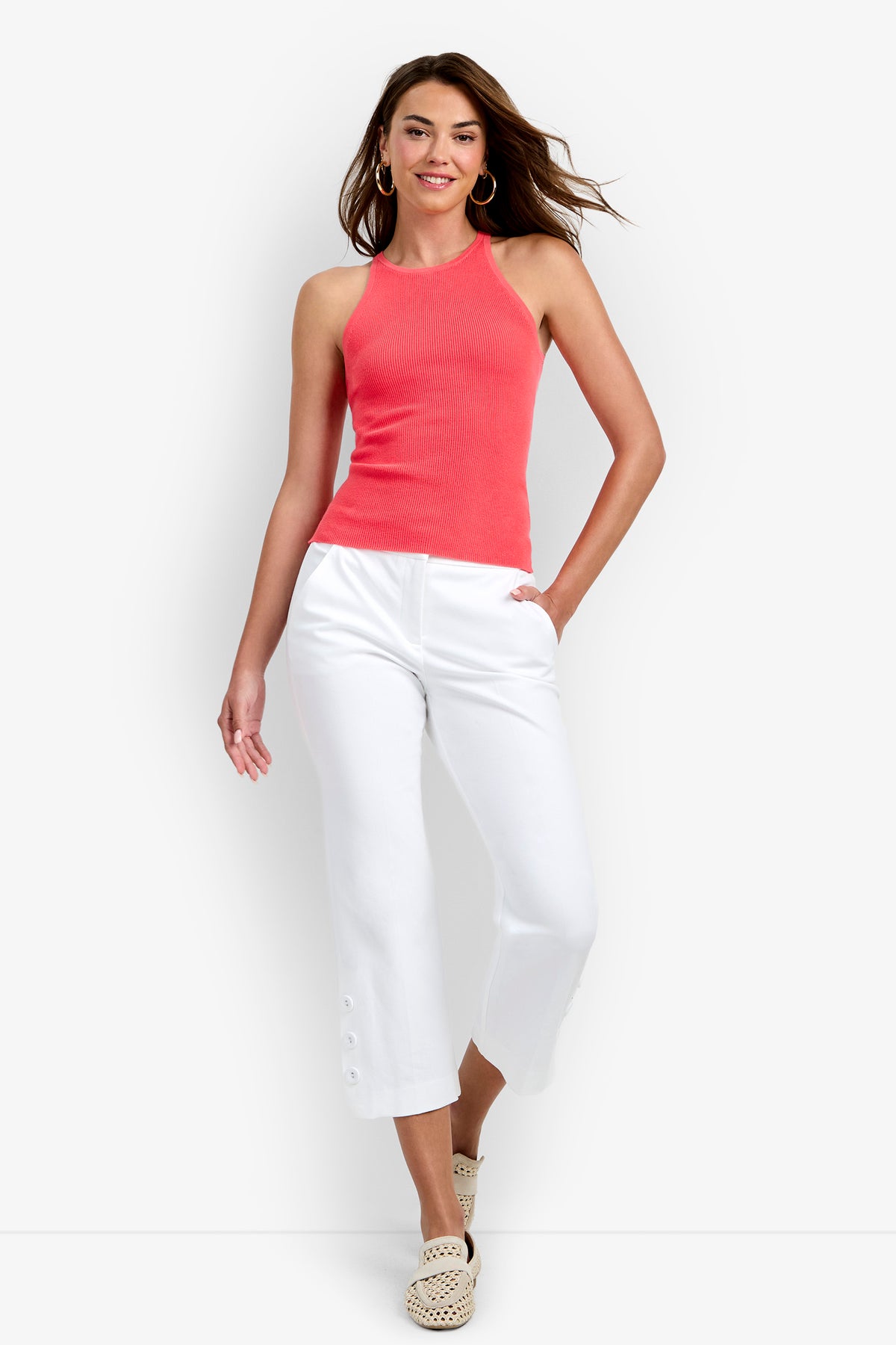 A woman wearing the Intercontinental Knit Tank in Corso Coral, paired with white cropped pants featuring buttons at the hem, hoop earrings, and woven slip-on shoes, stands and smiles against a plain white background.