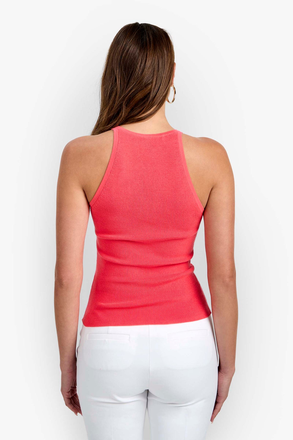 A woman with long brown hair is shown from the back against a plain light background, wearing the sleeveless coral Intercontinental Knit Tank and white pants.