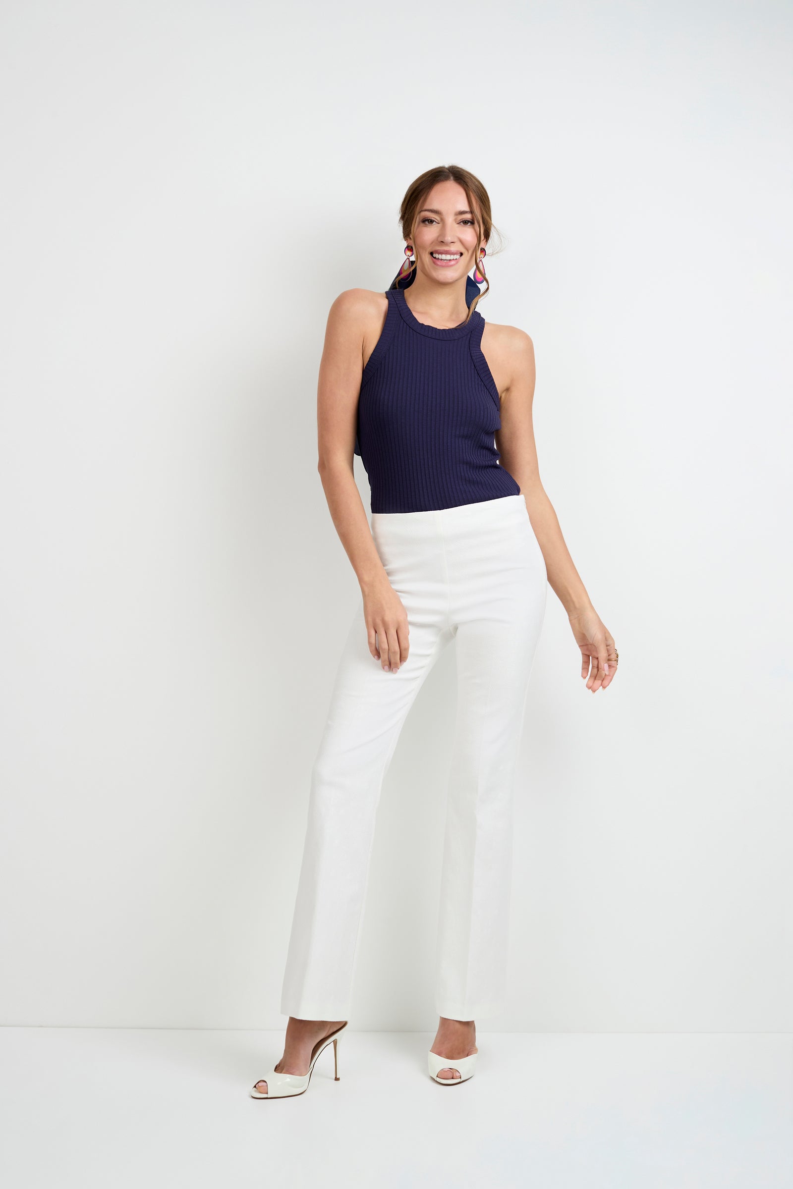 A woman stands against a plain white background, smiling. She wears the Continental Tank Top in navy blue, white high-waisted pants, white high-heeled shoes, and large pink earrings.