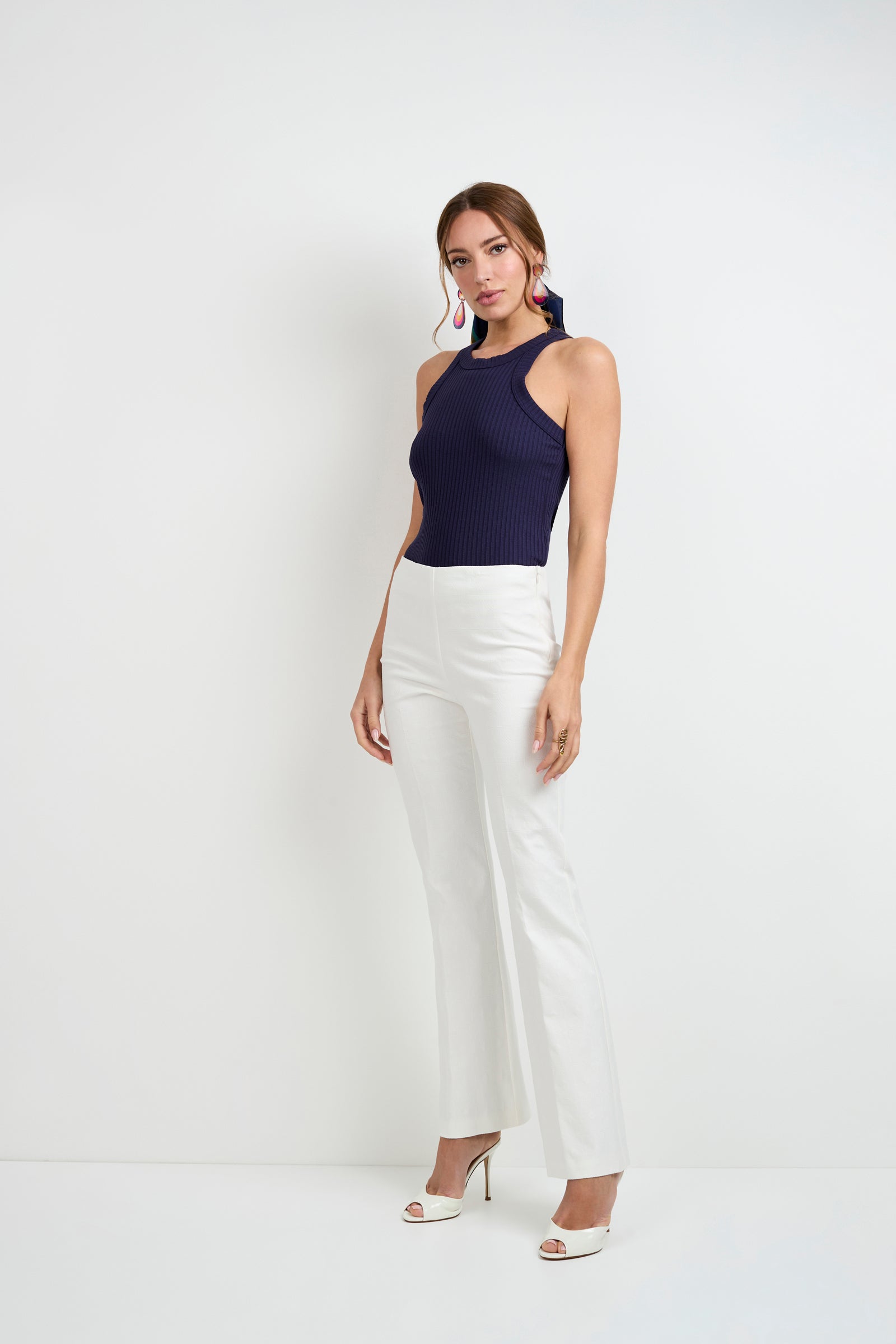 Woman with light brown hair pulled back poses against a white background, wearing the Continental Tank Top in navy, paired with white high-waisted pants, white open-toe heels, and colorful earrings. One hand rests by her side.