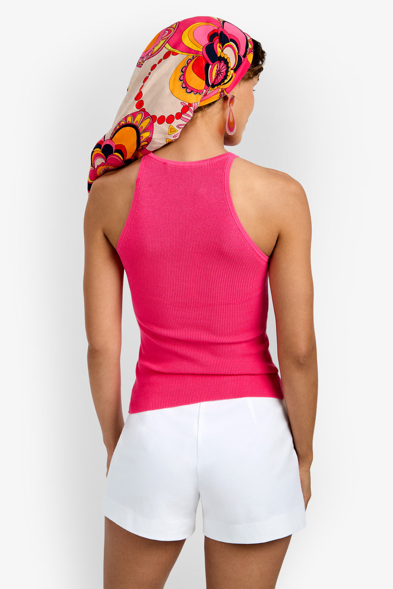A woman is seen from behind wearing the Intercontinental Knit Tank (Wholesale) in bright pink, paired with white shorts and a vibrant headscarf featuring pink, orange, and yellow patterns; she accessorizes with large hoop earrings.