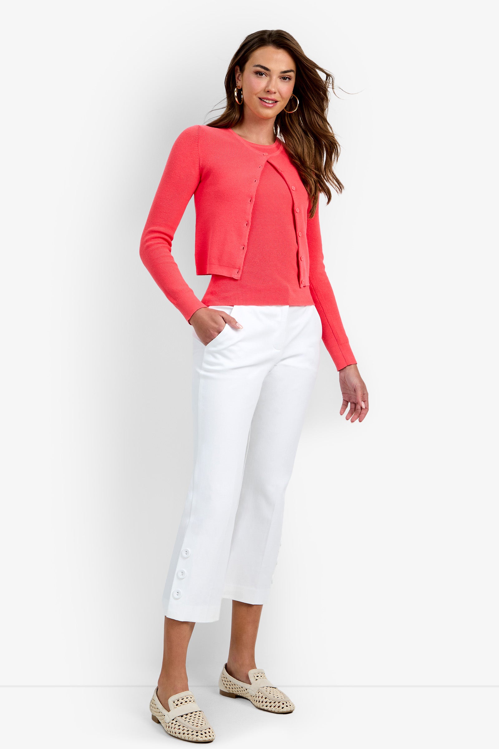 Certainly! Here’s a rewritten version using the product name: A woman stands smiling, wearing the Transcontinental Knit Cardigan in coral with a matching top, paired with white cropped pants featuring side buttons and cream woven loafers. She has long brown hair, hoop earrings, and one hand casually placed in her pocket.