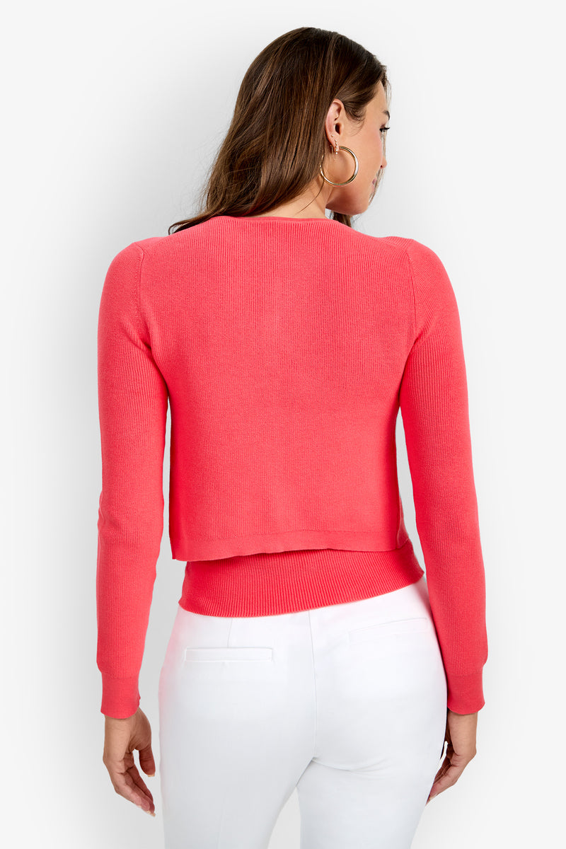 A woman with long brown hair, standing with her back to the camera against a plain white background, models the Transcontinental Knit Cardigan in a bright coral shade paired with white pants.