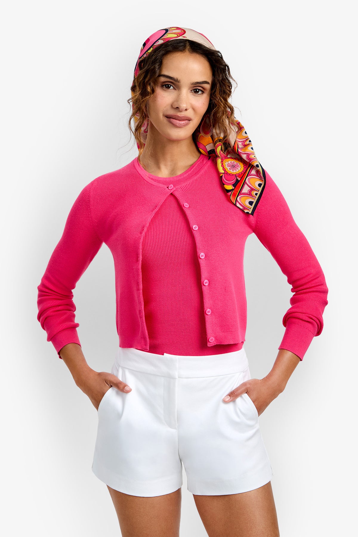 A woman wearing the Transcontinental Knit Cardigan in bright pink over a matching top, paired with white high-waisted shorts and a colorful patterned headscarf, stands with her hands on her hips against a plain white background.