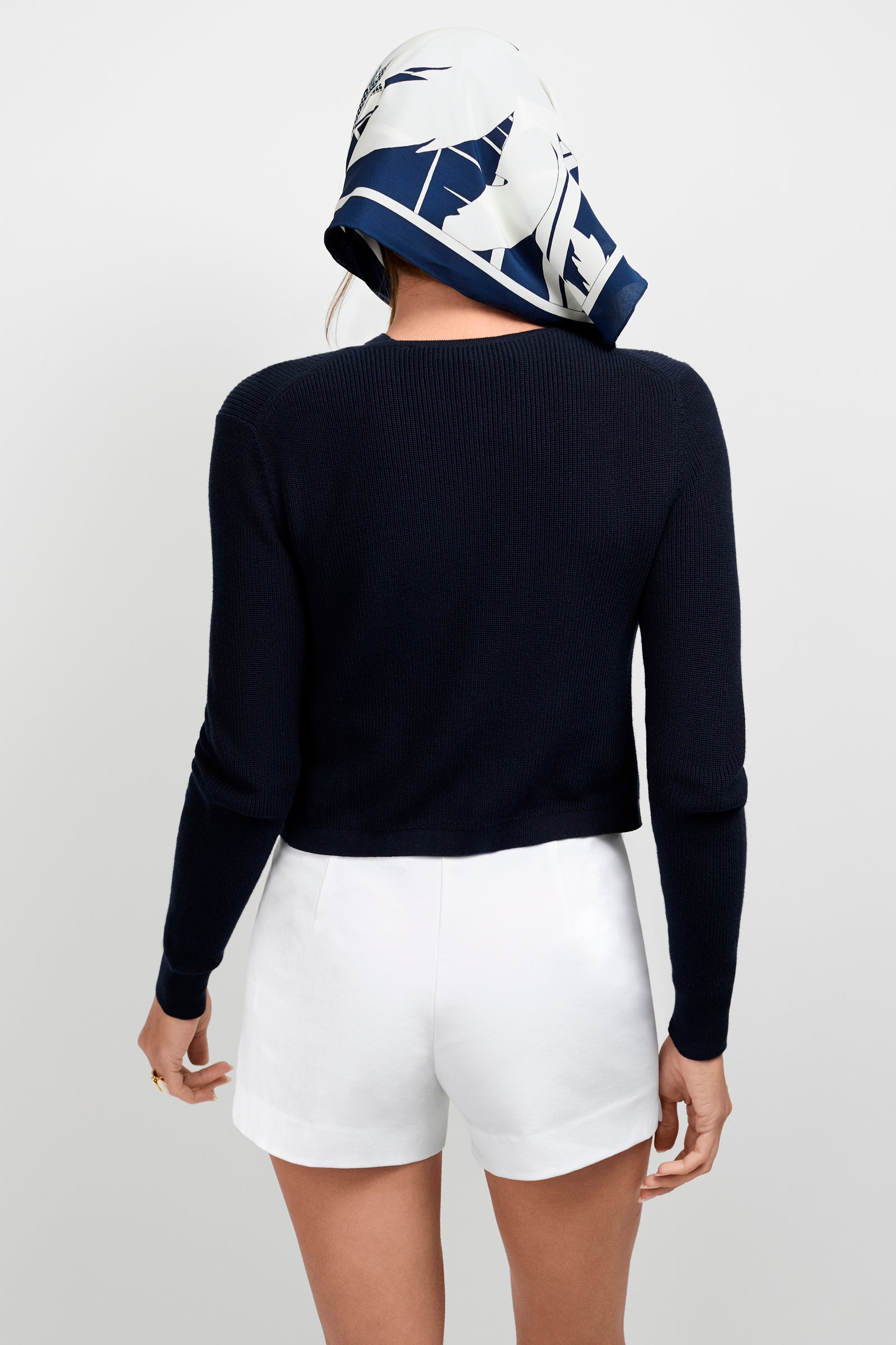 A person stands facing away, wearing the Transcontinental Knit Cardigan, white shorts, and a blue and white patterned headscarf against a plain light background.