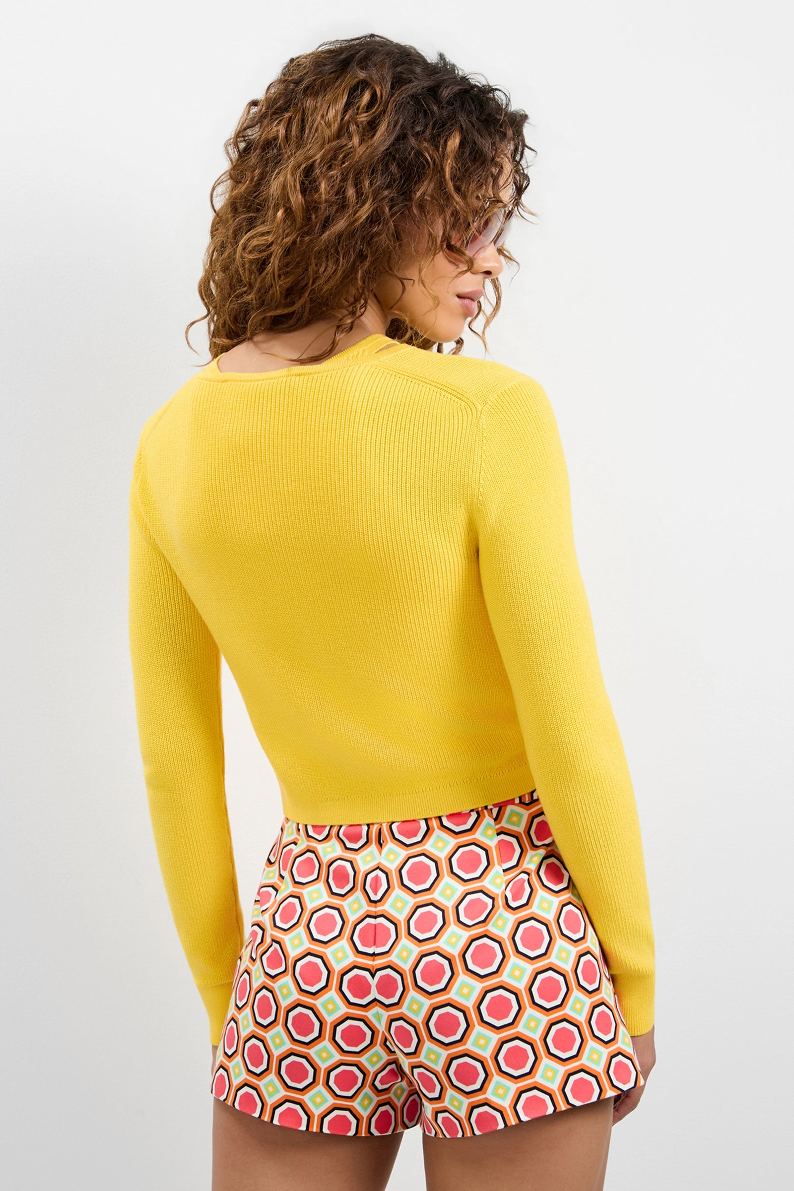 A woman with curly hair faces away, wearing the Transcontinental Knit Cardigan in bright yellow and high-waisted shorts with a red, yellow, and white geometric hexagon pattern. The background is plain white.