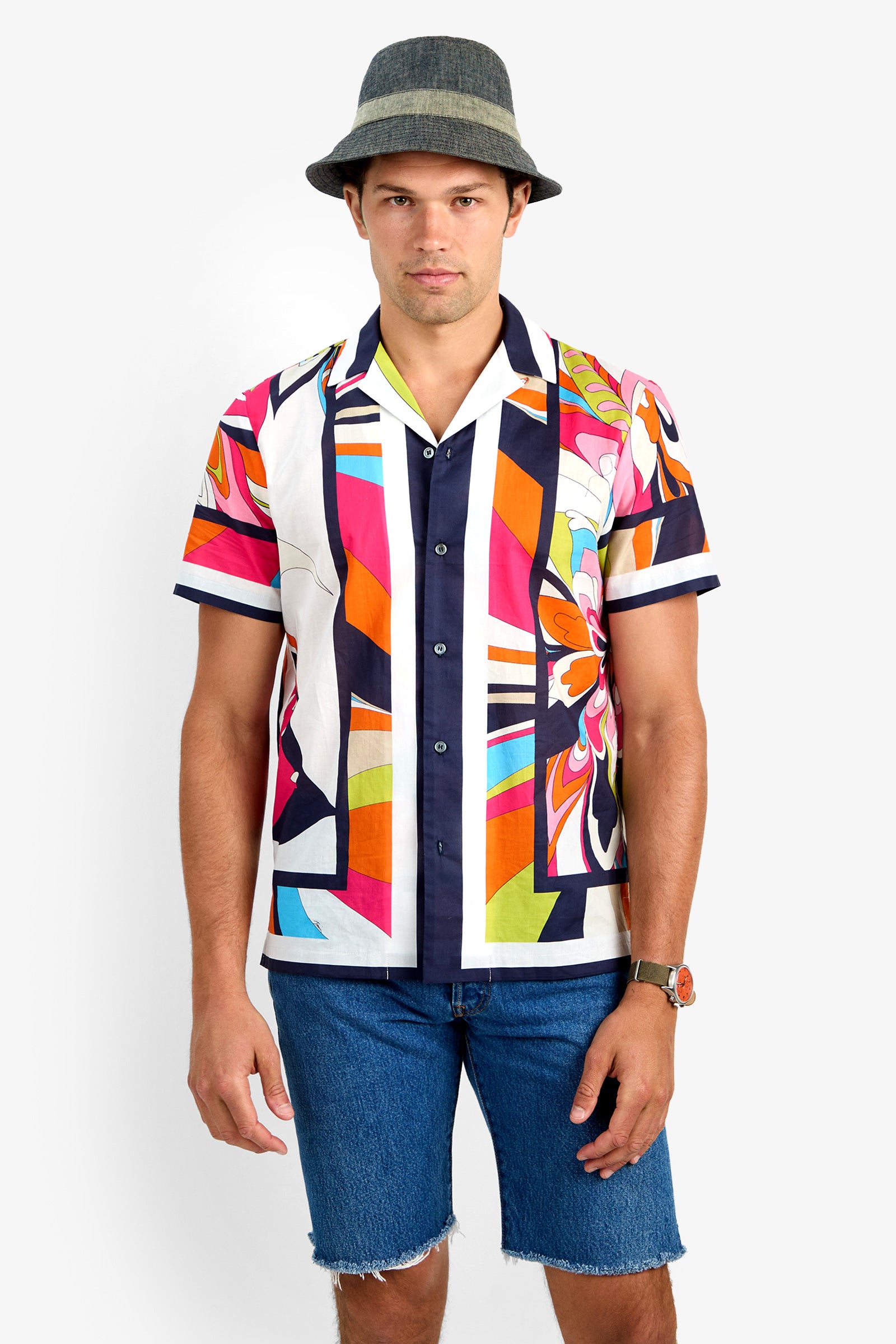 A man in a gray hat, the JOSUE CAMP SHIRT (100% cotton, colorful patterned short sleeves), blue denim shorts, and a watch stands facing the camera against a plain white background.