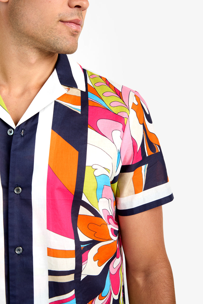 A man models the JOSUE CAMP SHIRT, a pure cotton, short-sleeve button-up with a bold abstract floral and geometric print in orange, pink, blue, green, and white. His upper body is pictured against a plain background.