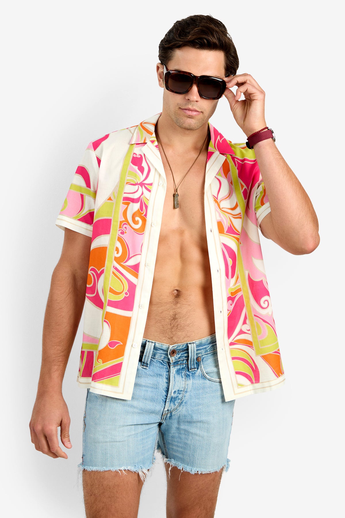 A man wearing dark sunglasses, the JOSUE CAMP SHIRT (a colorful patterned cotton short-sleeve shirt), denim shorts, and a necklace stands against a plain white background.