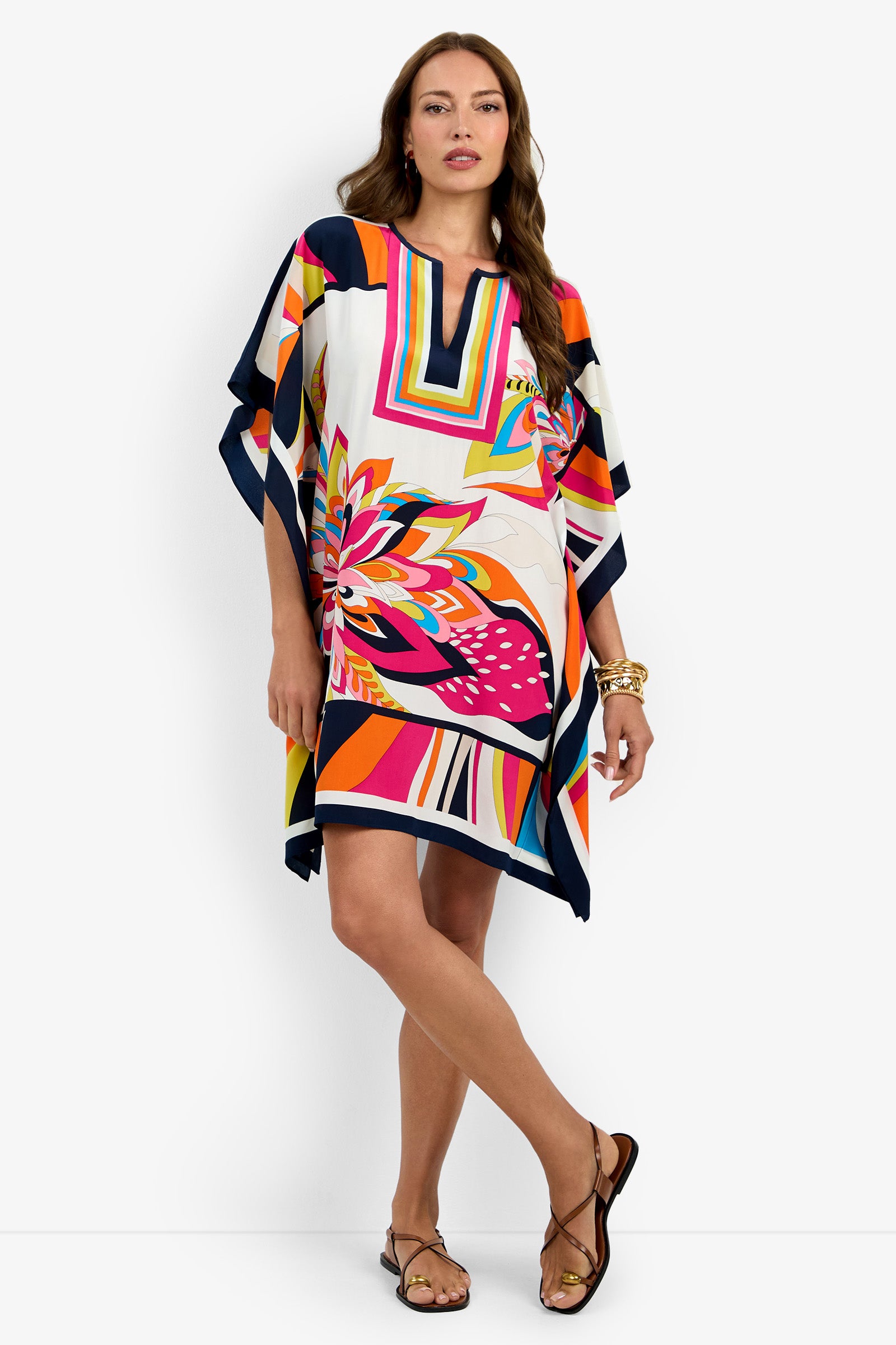 A woman in the Theodora Caftan Dress (Multi), featuring bold geometric and floral patterns, stands against a white background. She accessorizes with sandals and gold jewelry.