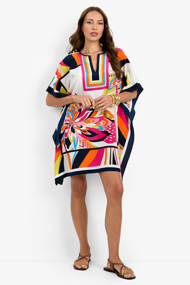 A woman stands against a plain white background wearing Theodora Caftan Dress, featuring a colorful geometric and floral pattern, styled with gold bracelets and brown sandals.