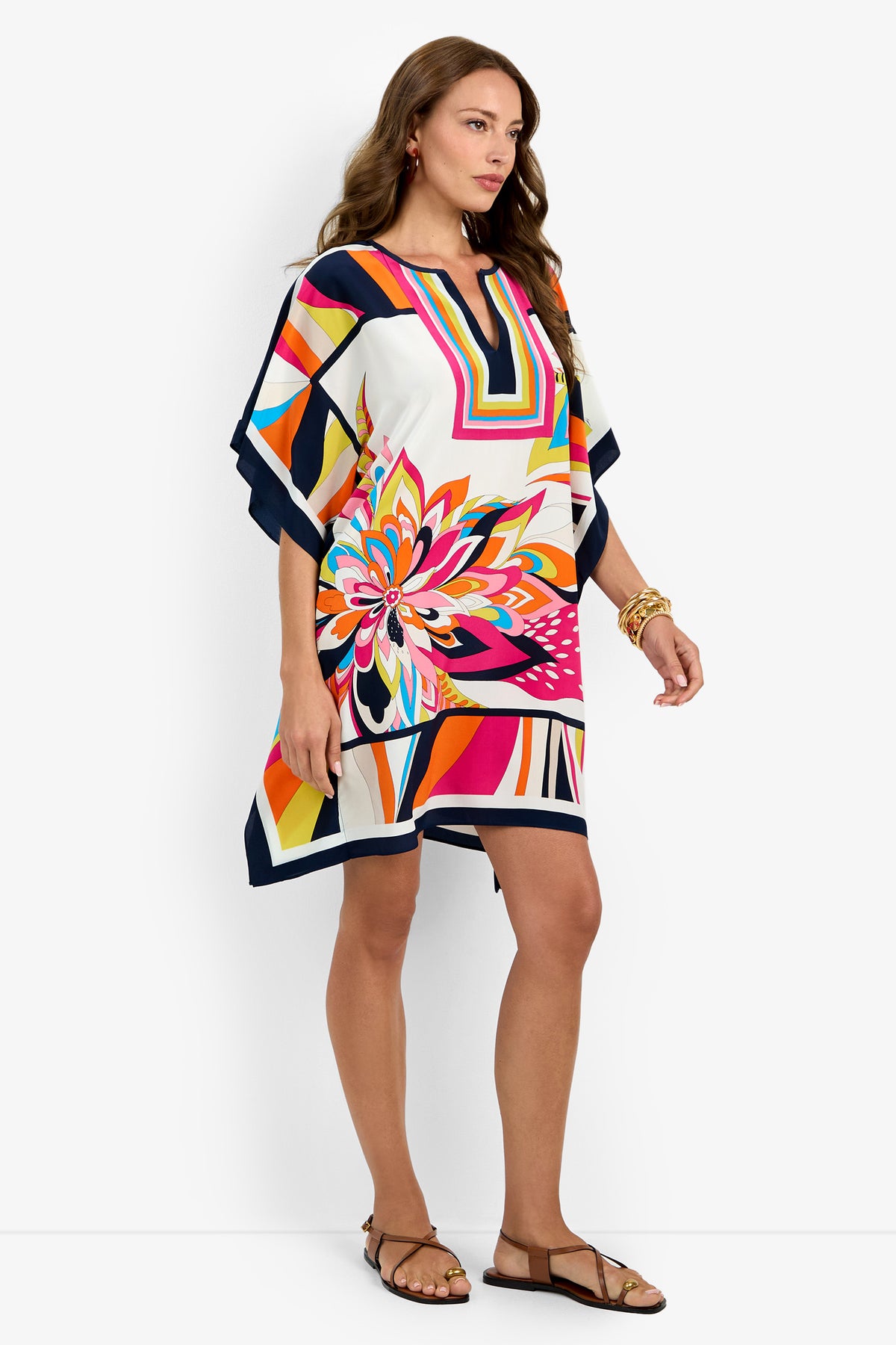 A woman stands sideways in Theodora Caftan Dress, featuring colorful geometric and floral patterns with a V-neckline, styled with sandals and gold bracelets against a plain white background.