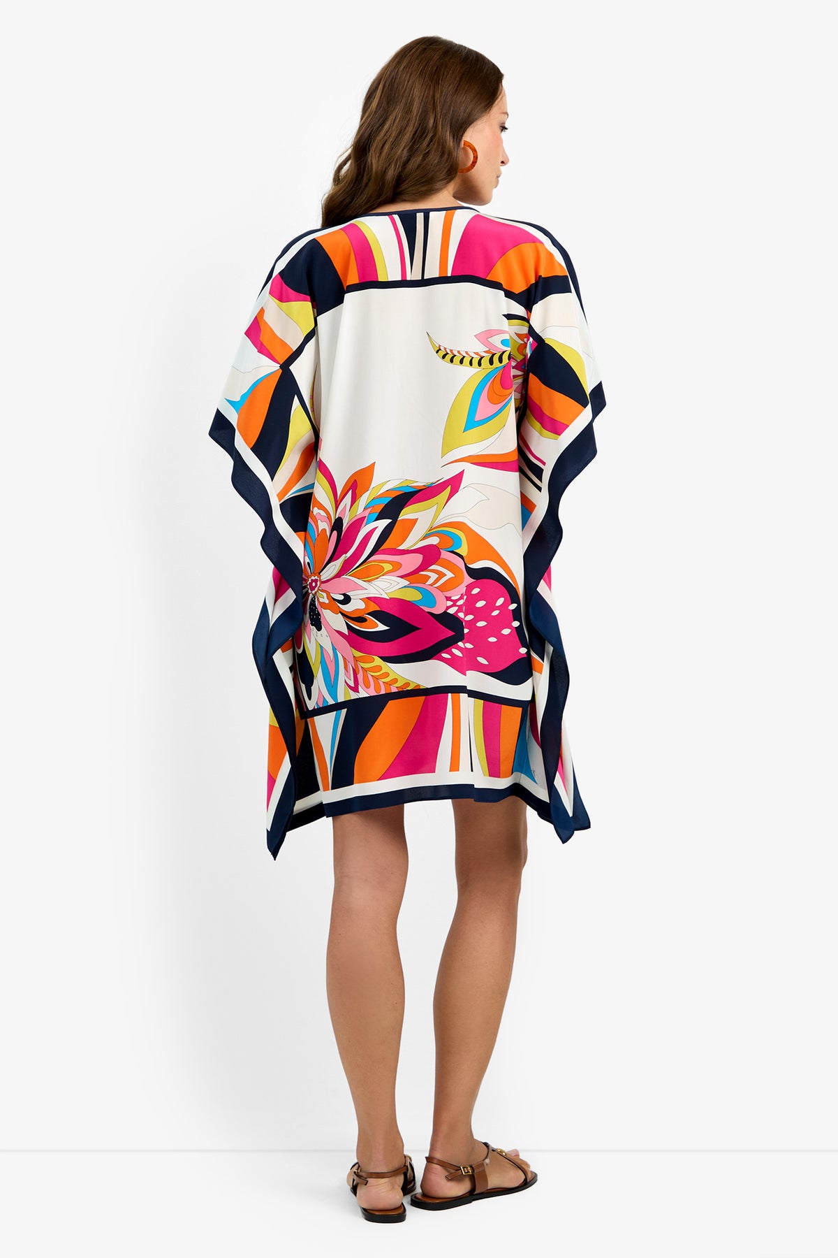 A woman with brown hair stands facing away in Theodora Caftan Dress—colorful, loose-fitting with vibrant abstract patterns—and flat sandals, against a plain white background.