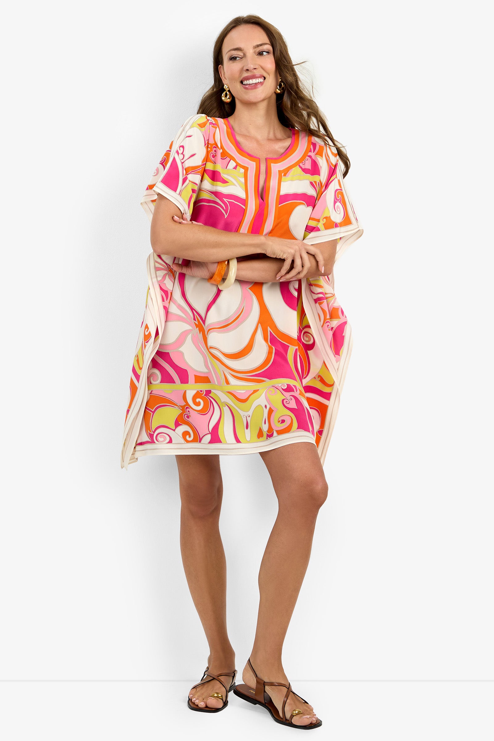 A woman wearing the Theodora Caftan Dress, featuring pink, orange, and cream swirls, smiles against a white background. She pairs it with gold hoop earrings and sandals, her long hair flowing freely.
