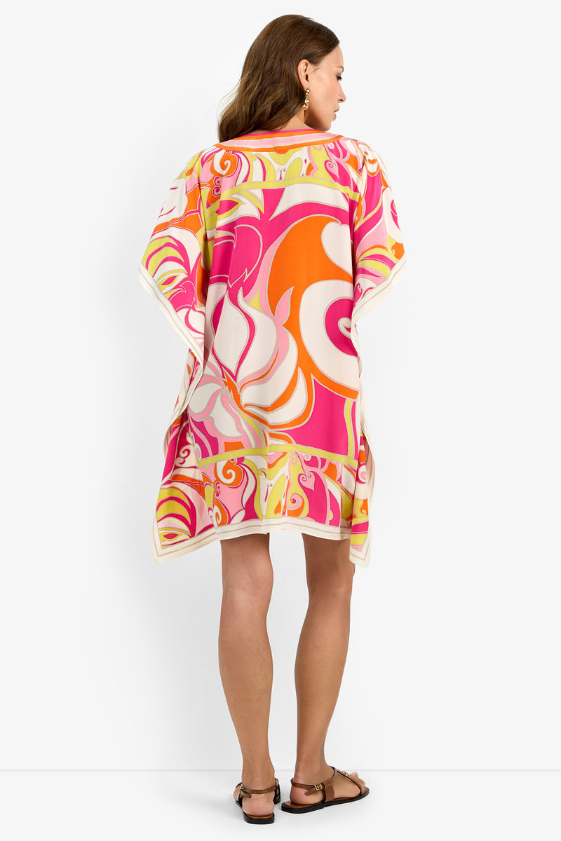 A woman with long brown hair stands with her back to the camera, wearing the Theodora Caftan Dress— a colorful, loose-fitting tunic featuring bright orange, pink, yellow, and white swirling patterns. She pairs it with flat sandals.