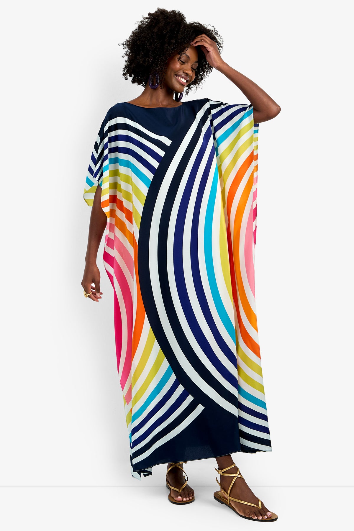 A smiling woman wears the Theodora Caftan Maxi Dress, a long pure silk dress with bold curved stripes in navy, white, pink, orange, yellow, and blue. She stands against a plain white background with one hand touching her hair.