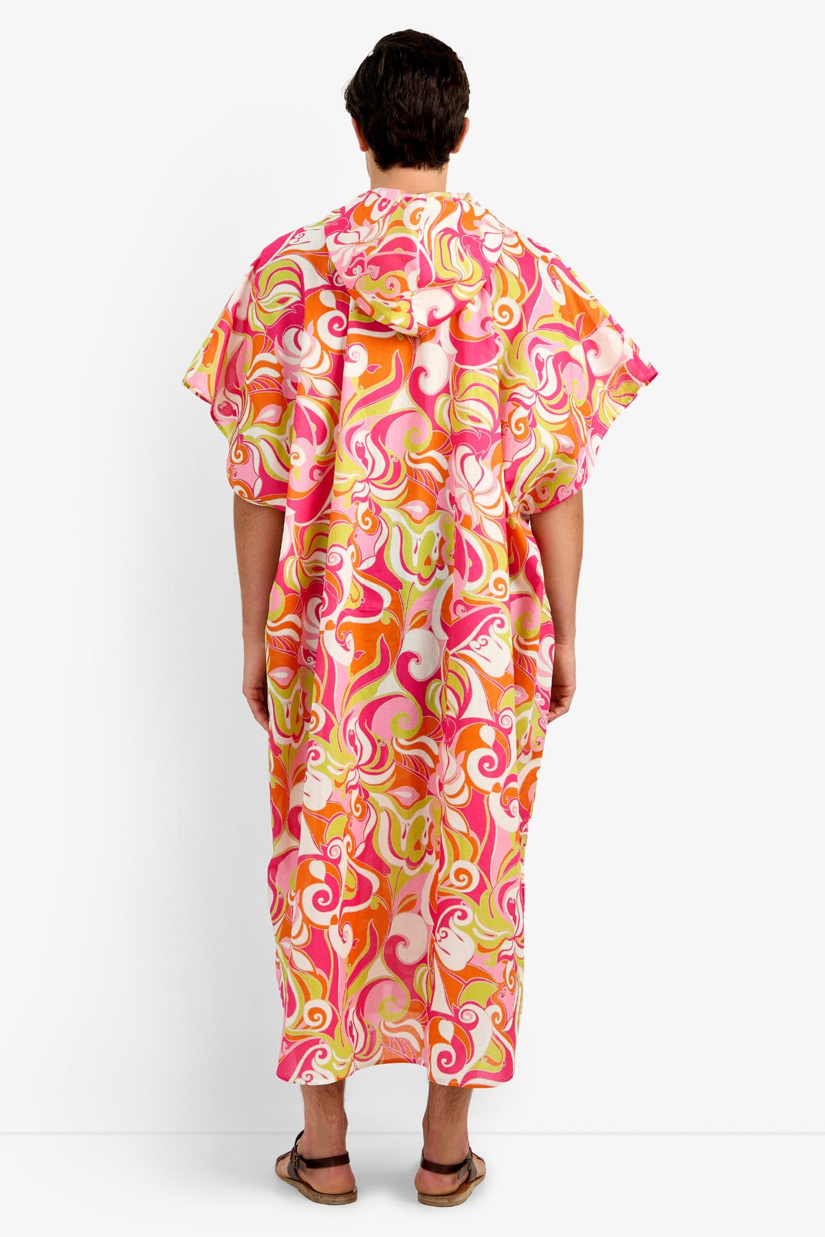 A person stands against a white background wearing the HURLEY CAFTAN, a loose, hooded 100% cotton garment with wide sleeves in a vibrant pink, orange, and yellow swirl pattern. They are also wearing sandals.