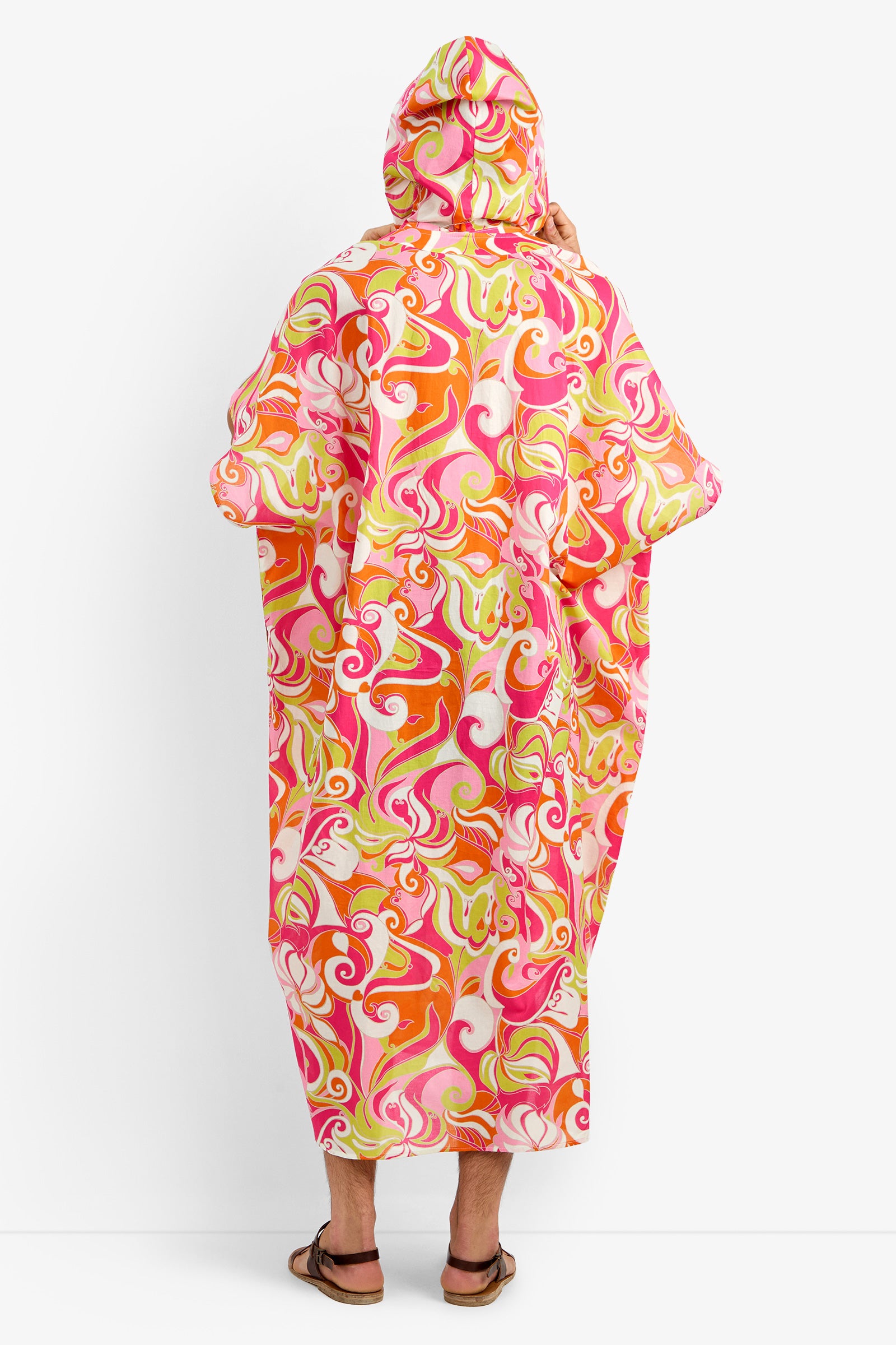 A person stands with their back turned, wearing the HURLEY CAFTAN—a 100% cotton hooded poncho featuring a colorful swirl pattern in pink, orange, yellow, and white—paired with sandals against a plain white background.