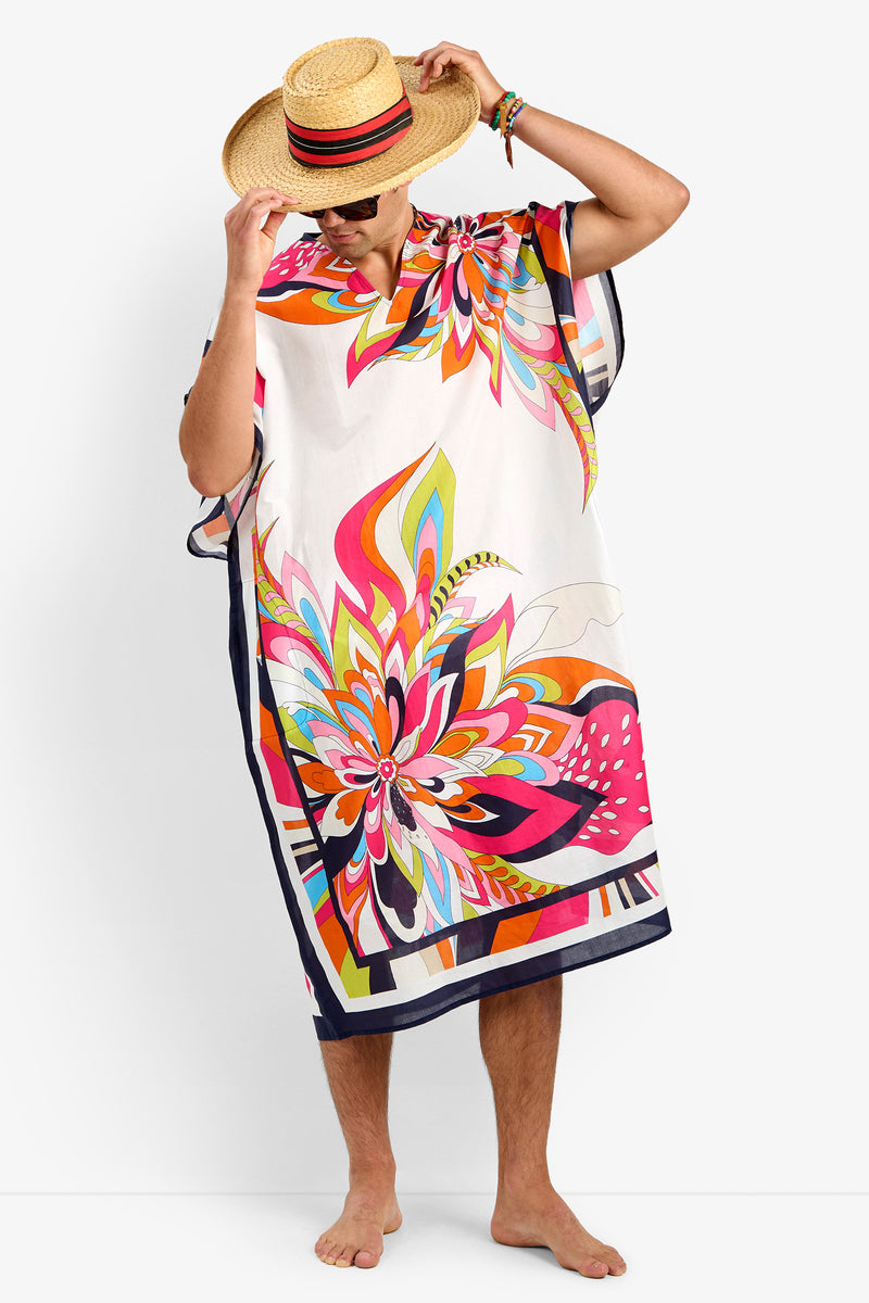 A person in the ST. MARTIN kaftan (MULTI), 100% cotton with a floral print, and a straw hat stands barefoot on a plain white background, tilting the hat to partially cover their face.