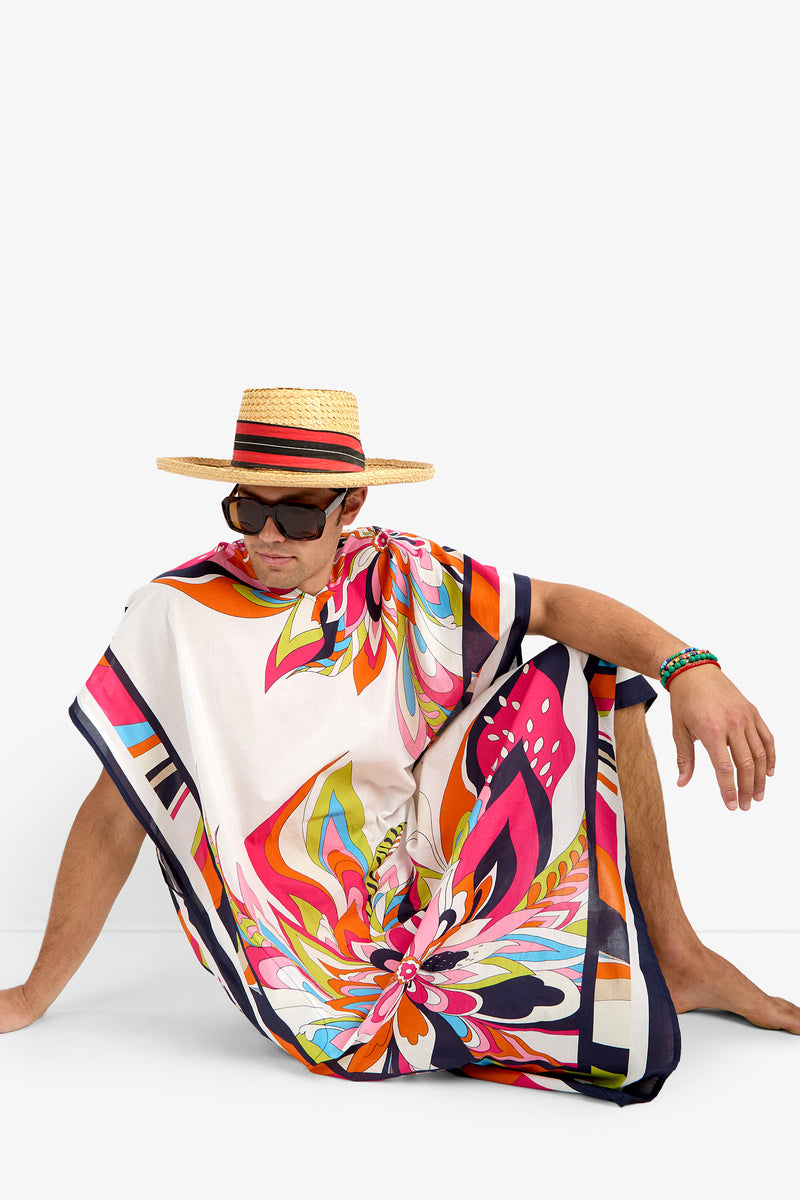 A man in the ST. MARTIN colorful patterned kaftan, soft cotton fabric, straw hat with striped band, and large sunglasses sits on the floor against a plain white background.