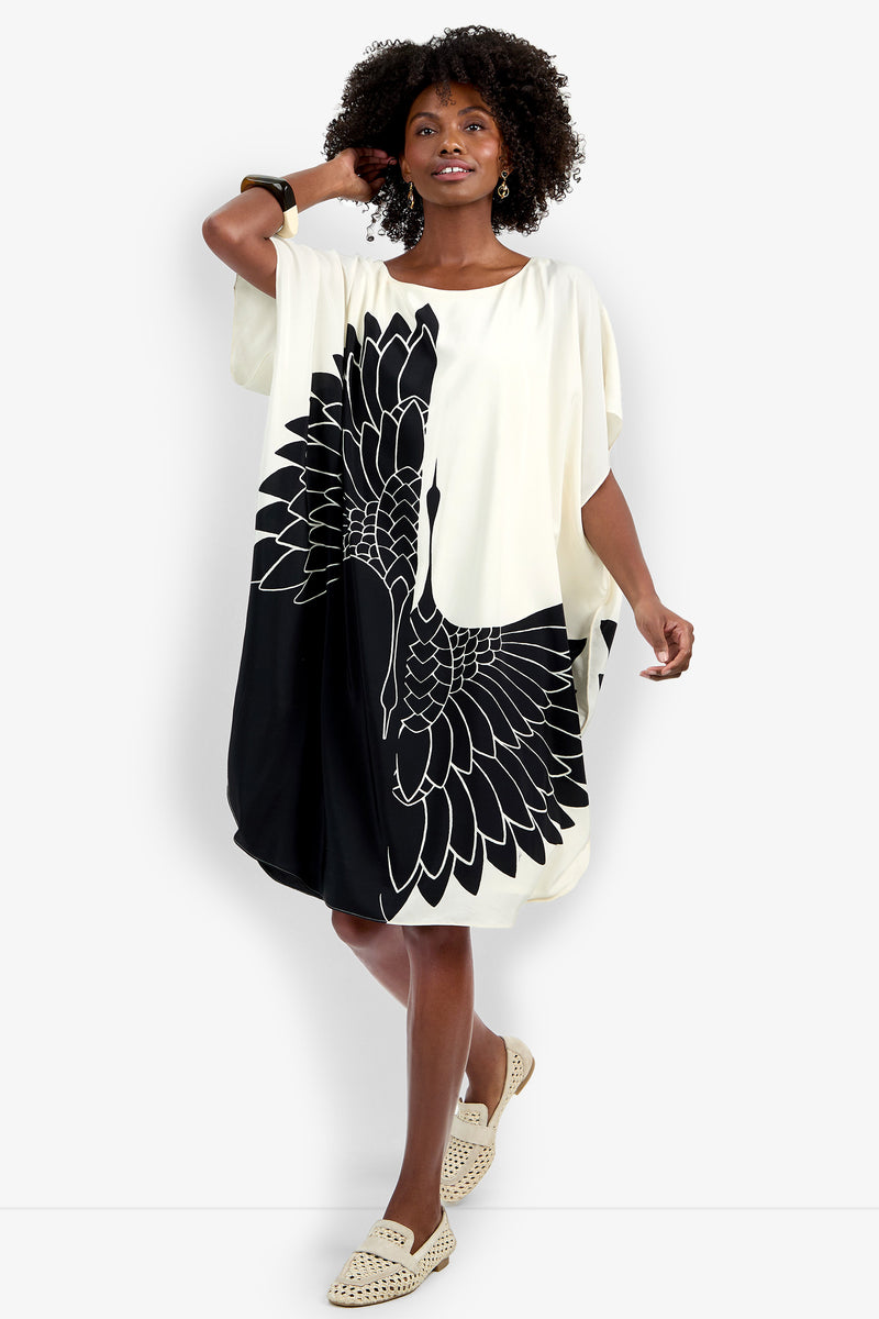 A woman models the Vintage Crane Caftan, a loose knee-length dress with a bold black crane print, one black sleeve and one white sleeve. She poses with an arm raised, wearing neutral shoes and a bangle.