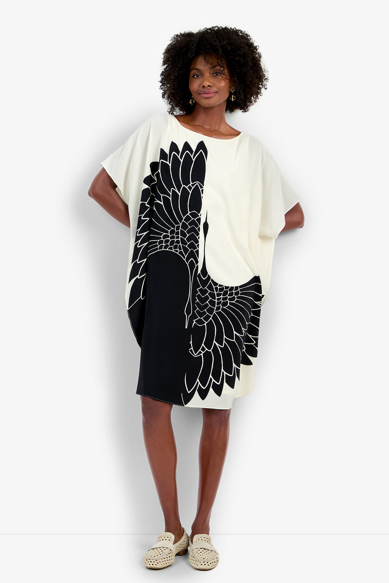 A woman with curly hair stands against a white background wearing the Vintage Crane Caftan, a loose cream short-sleeve dress with a bold black crane design, paired with woven beige slip-on shoes.