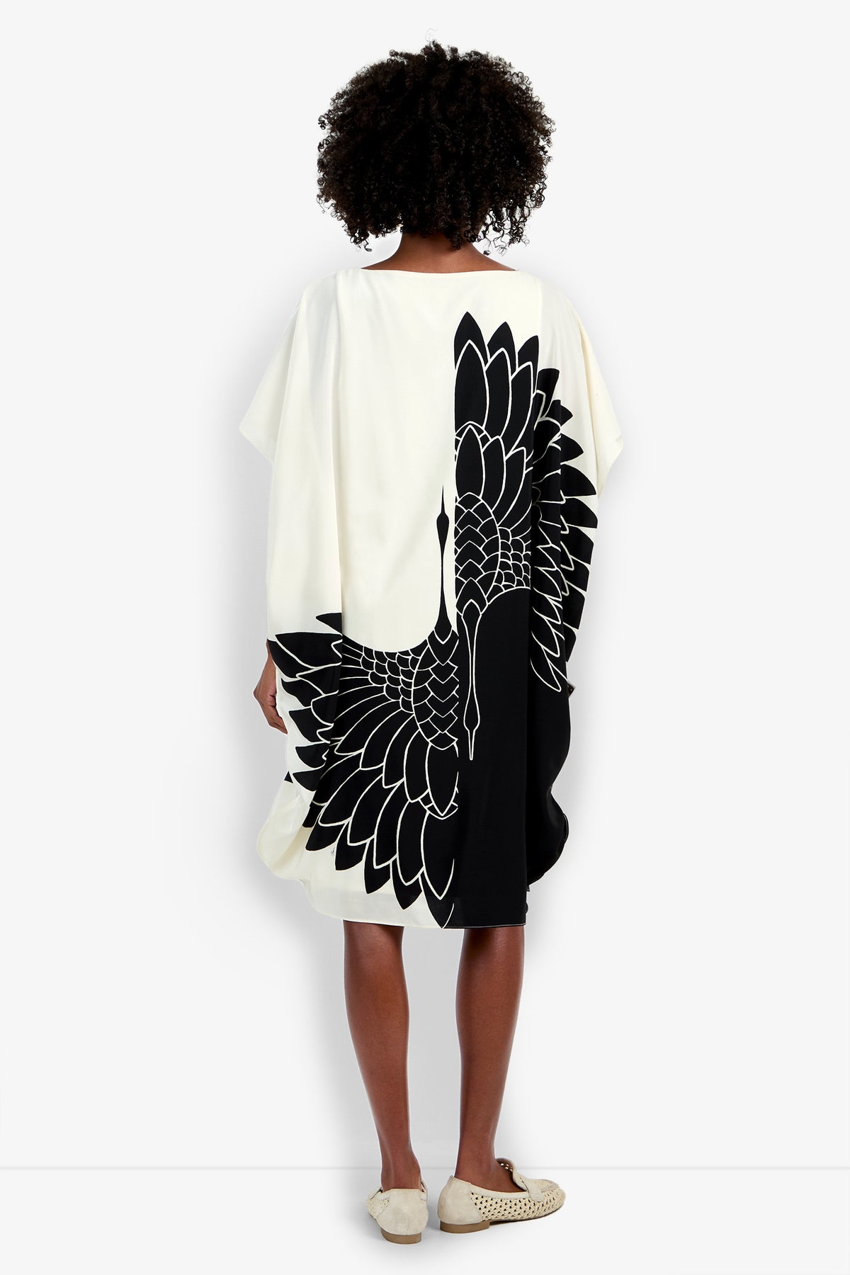 A person with curly hair faces away, wearing the Vintage Crane Caftan—a loose, knee-length white dress featuring a bold black stylized crane on the back—paired with light shoes.