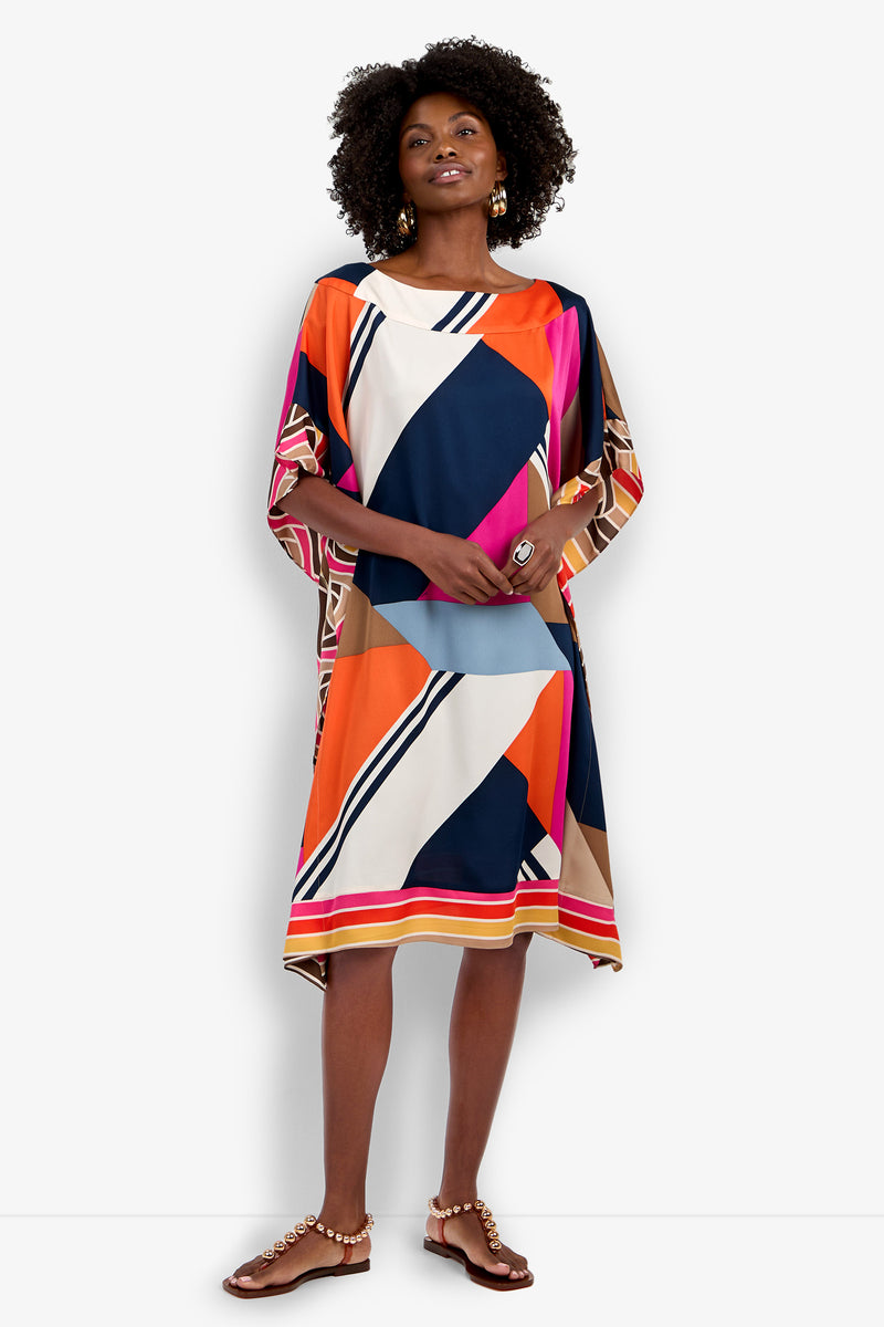 A smiling woman wears the Vintage Geo Caftan, a loose, knee-length dress with bold orange, navy, pink, blue, cream, and beige geometric patterns. She stands with hands in pockets, styled with sandals and earrings.