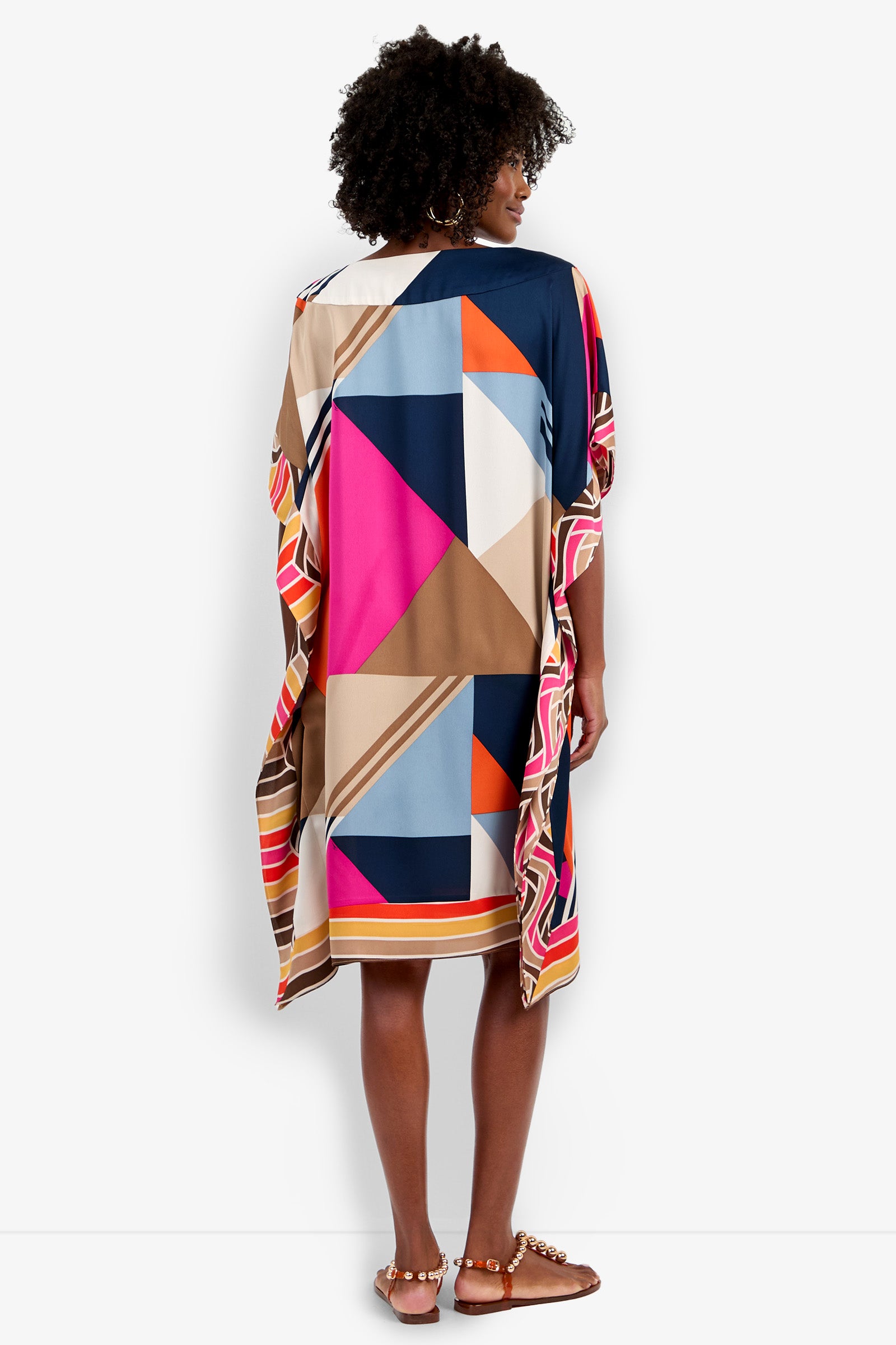 A woman with curly hair faces away, wearing the Vintage Geo Caftan—a loose, knee-length dress featuring bold blue, pink, tan, and white geometric patterns—paired with gold-detailed flat sandals.