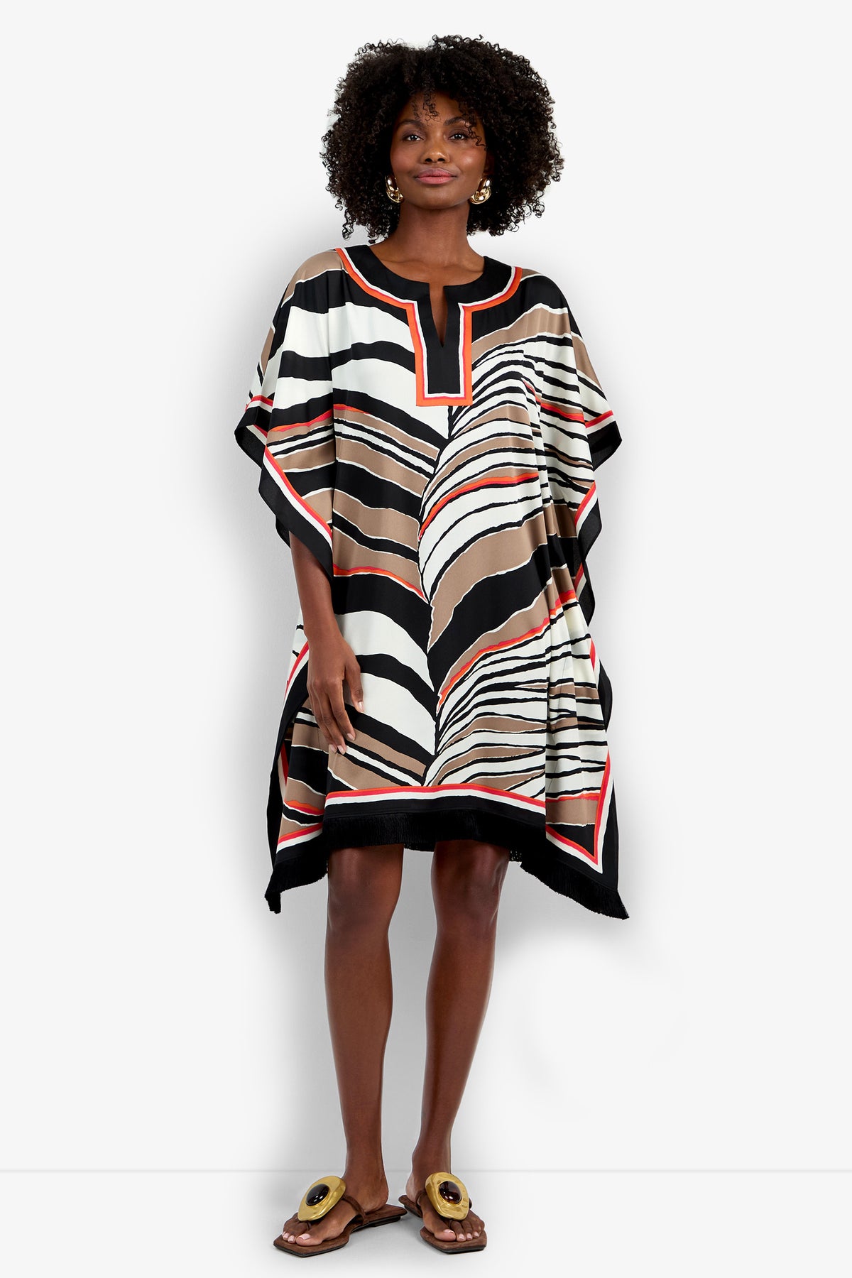 A woman with curly hair stands on a white background, wearing the Vintage Zebra Caftan—a loose, short dress featuring bold brown, black, white, and orange abstract stripes. She pairs it with gold-accented sandals.