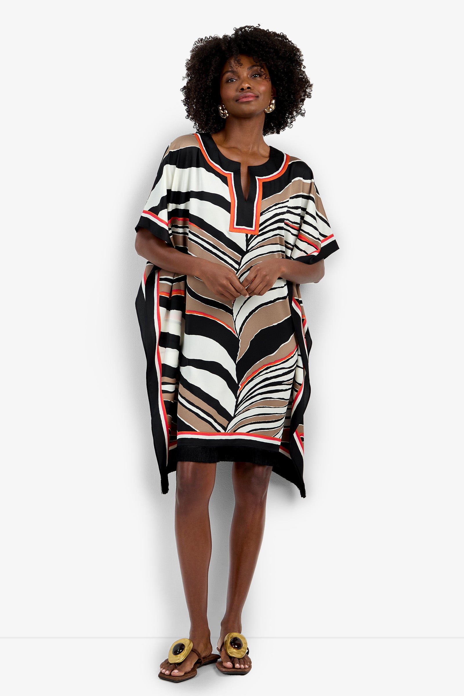 A woman with curly hair and large earrings wears the Vintage Zebra Caftan, a loose, short-sleeved dress featuring an abstract black, white, beige, and red pattern. She pairs it with flat sandals accented in gold.