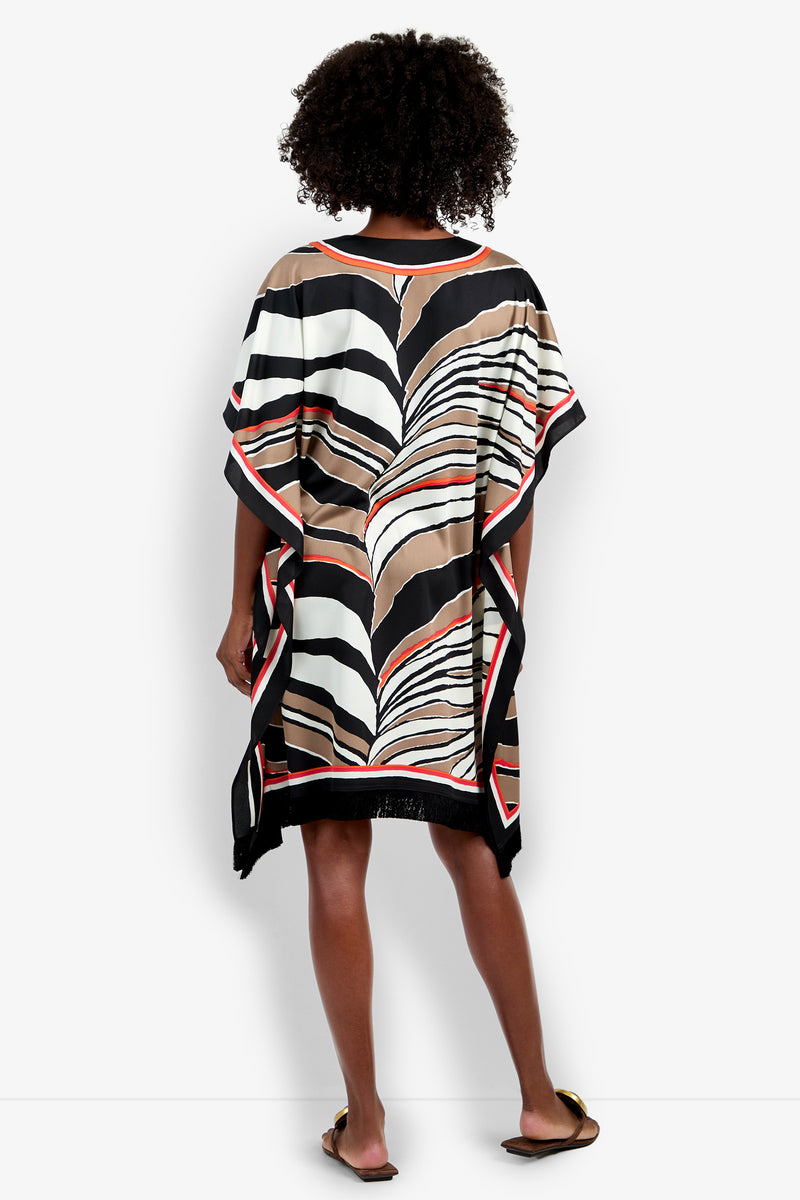A person with curly hair stands facing away, wearing the Vintage Zebra Caftan—a loose, knee-length dress with bold black, white, brown, and red abstract stripes—paired with flat sandals against a plain background.