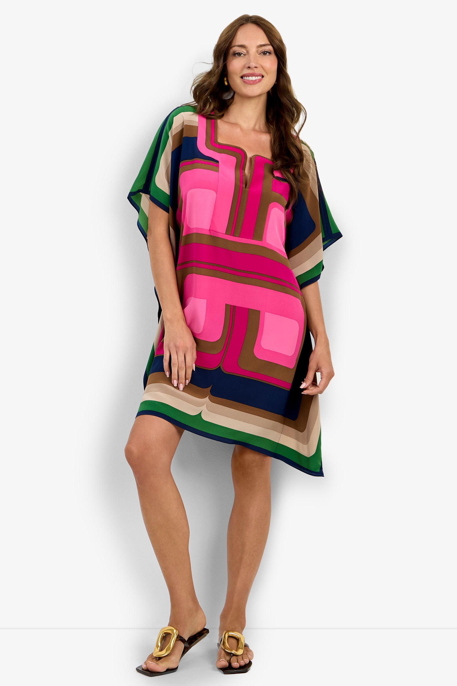 A woman wears the Vintage dress—a loose, colorful piece with geometric patterns in pink, green, blue, and brown—smiling as she stands with one hand on her hip and the other relaxed by her side.