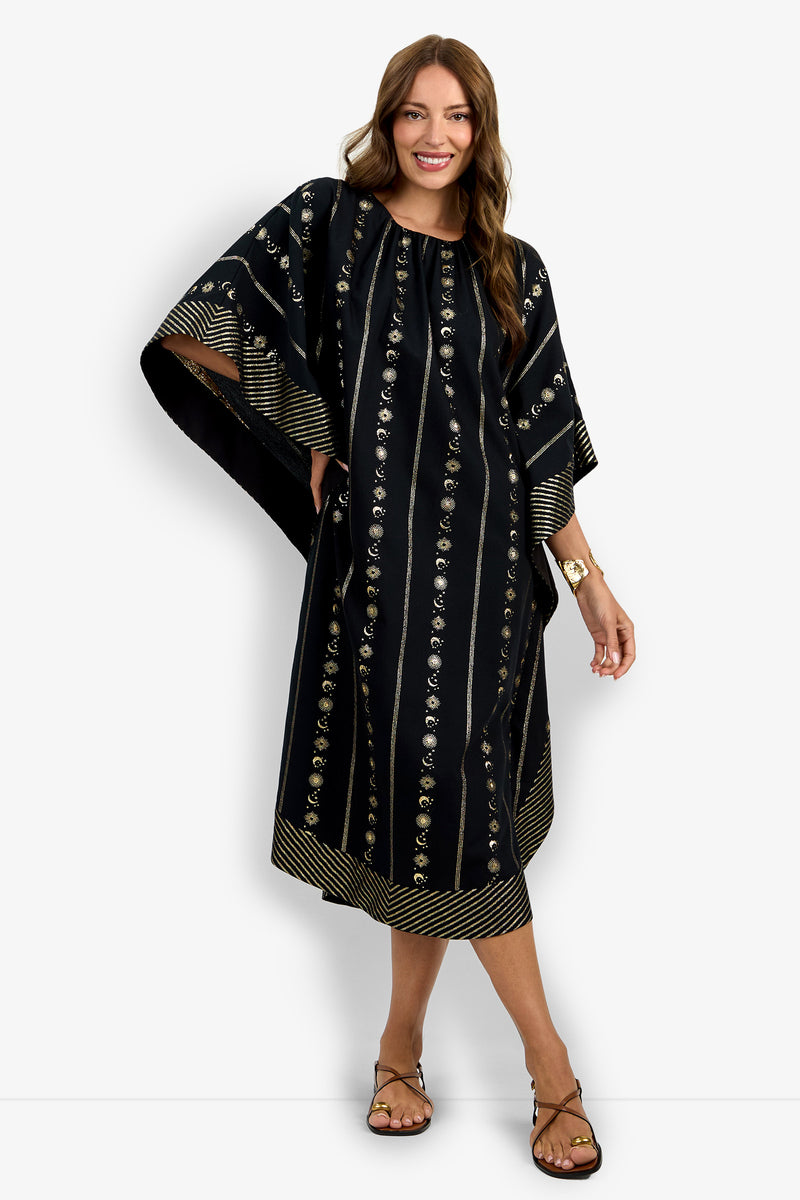 A woman with long brown hair smiles as she stands barefoot, wearing the Vintage Celestial Caftan—a loose, knee-length black caftan featuring gold stripes and decorative patterns—posing against a plain white background.
