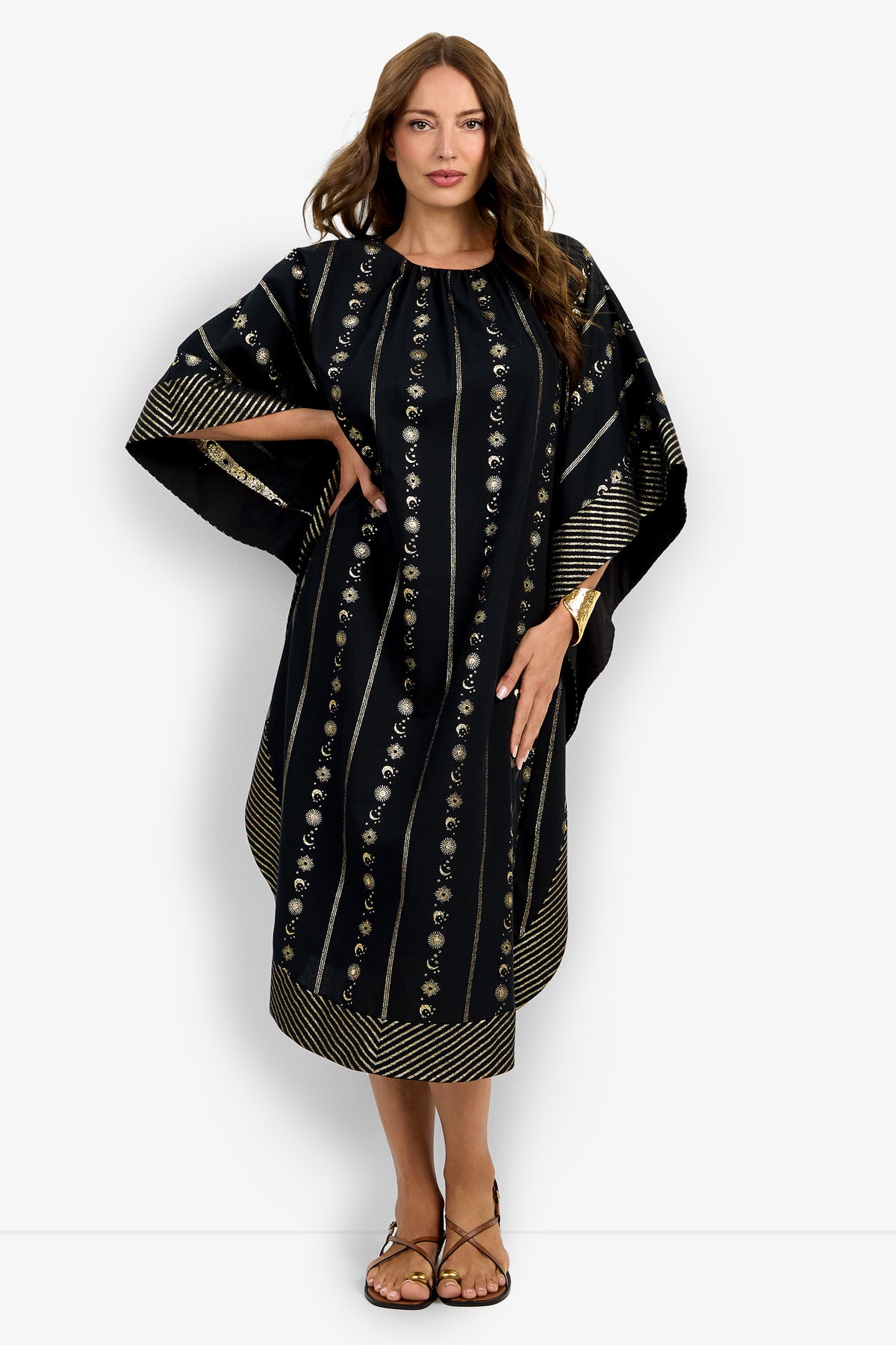 A woman wears the Vintage Celestial Caftan, a loose black dress with gold vertical stripes and floral patterns, featuring wide sleeves and a below-the-knee length. She has long brown hair, sandals, and a bracelet.