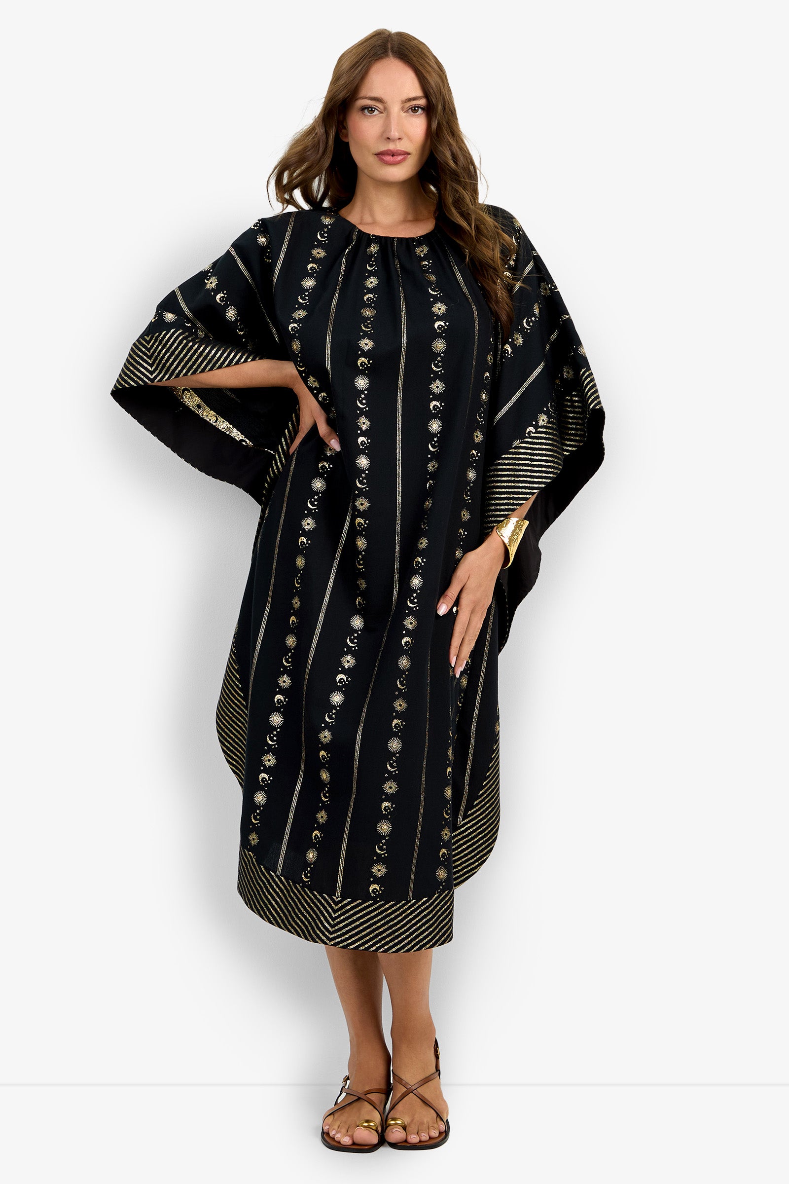 A woman wears the Vintage Celestial Caftan, a loose black dress with gold vertical stripes and floral patterns, featuring wide sleeves and a below-the-knee length. She has long brown hair, sandals, and a bracelet.