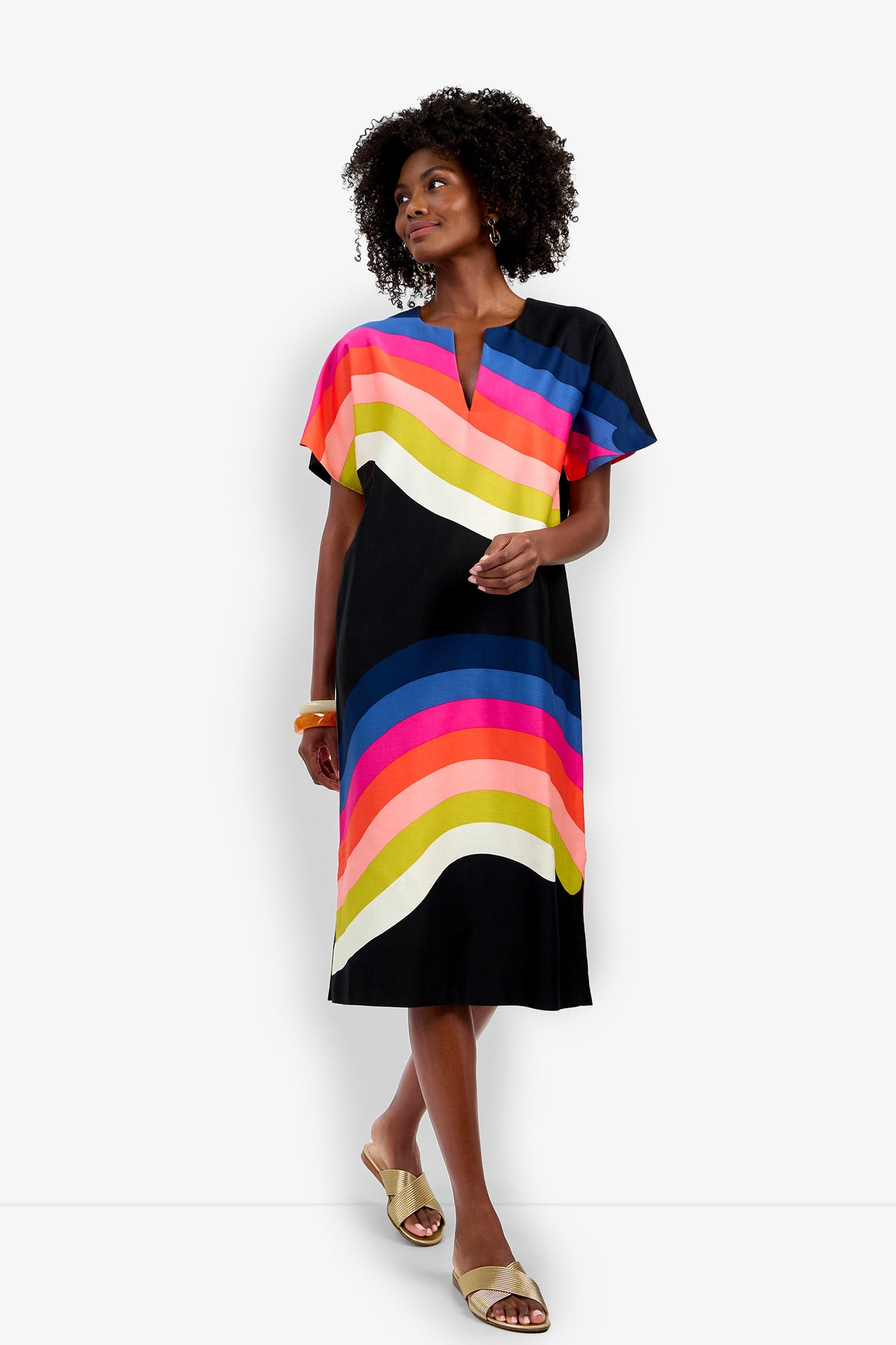 A woman models the Vintage Rainbow Caftan, a knee-length black dress with vibrant wavy stripes in yellow, pink, red, blue, and white. She accessorizes with tan sandals and orange bangles against a plain white background.