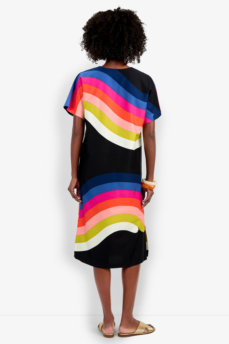A person with curly hair stands barefoot, seen from behind, wearing the Vintage Rainbow Caftan—a black knee-length dress featuring bold curved stripes in pink, orange, yellow, blue, and white—and holding bangle bracelets.