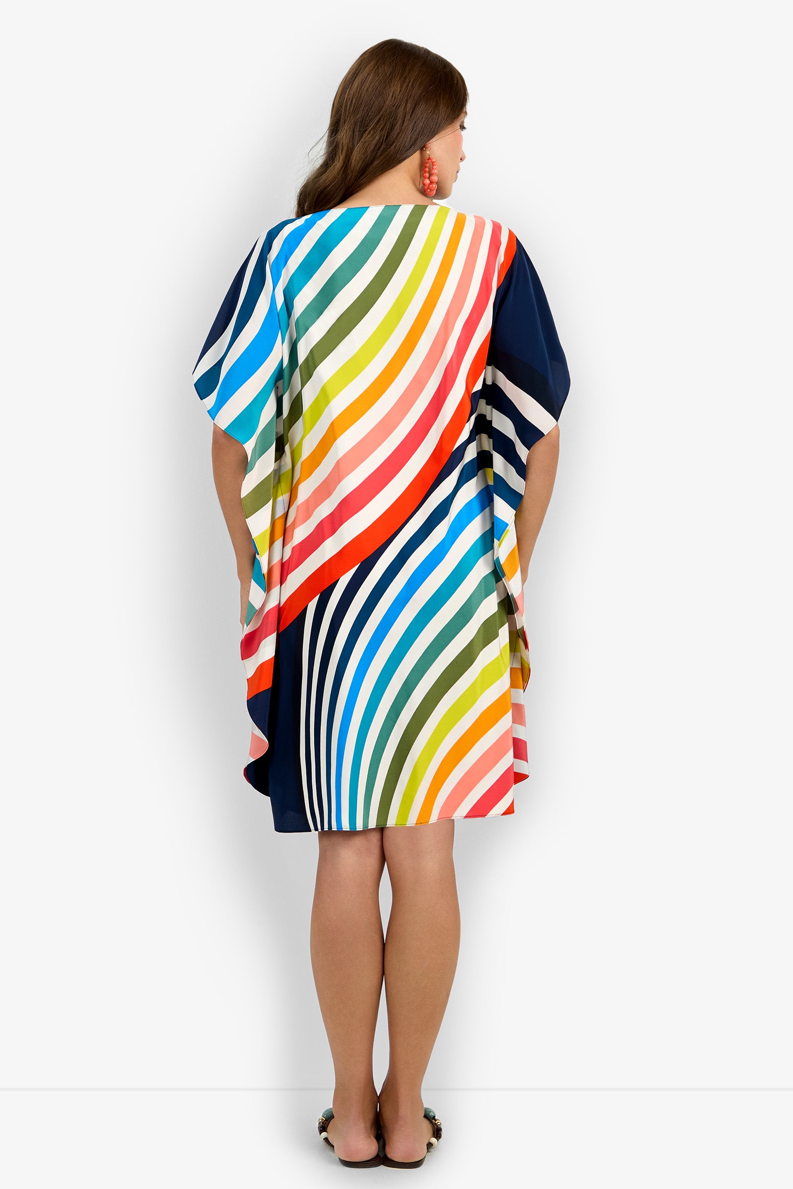 A woman with long brown hair stands facing away, wearing the Vintage Ombre Rainbow Caftan—a loose-fitting dress featuring colorful diagonal and curved stripes. She is also wearing sandals.