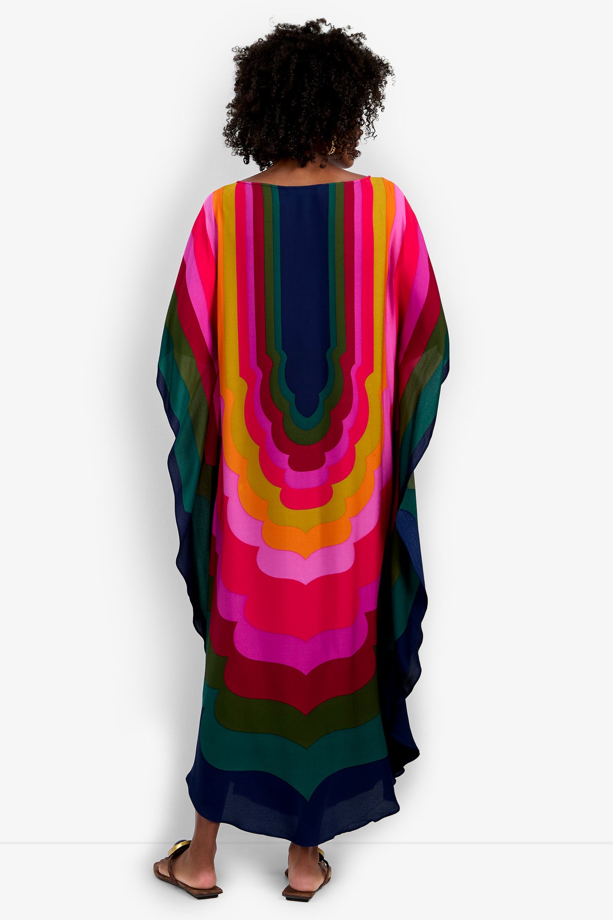Someone stands facing away in a Vintage Moroccan Geo Caftan, featuring bold, layered geometric patterns in pink, orange, yellow, green, and navy on white. The flowing ankle-length hem pairs perfectly with their sandals.