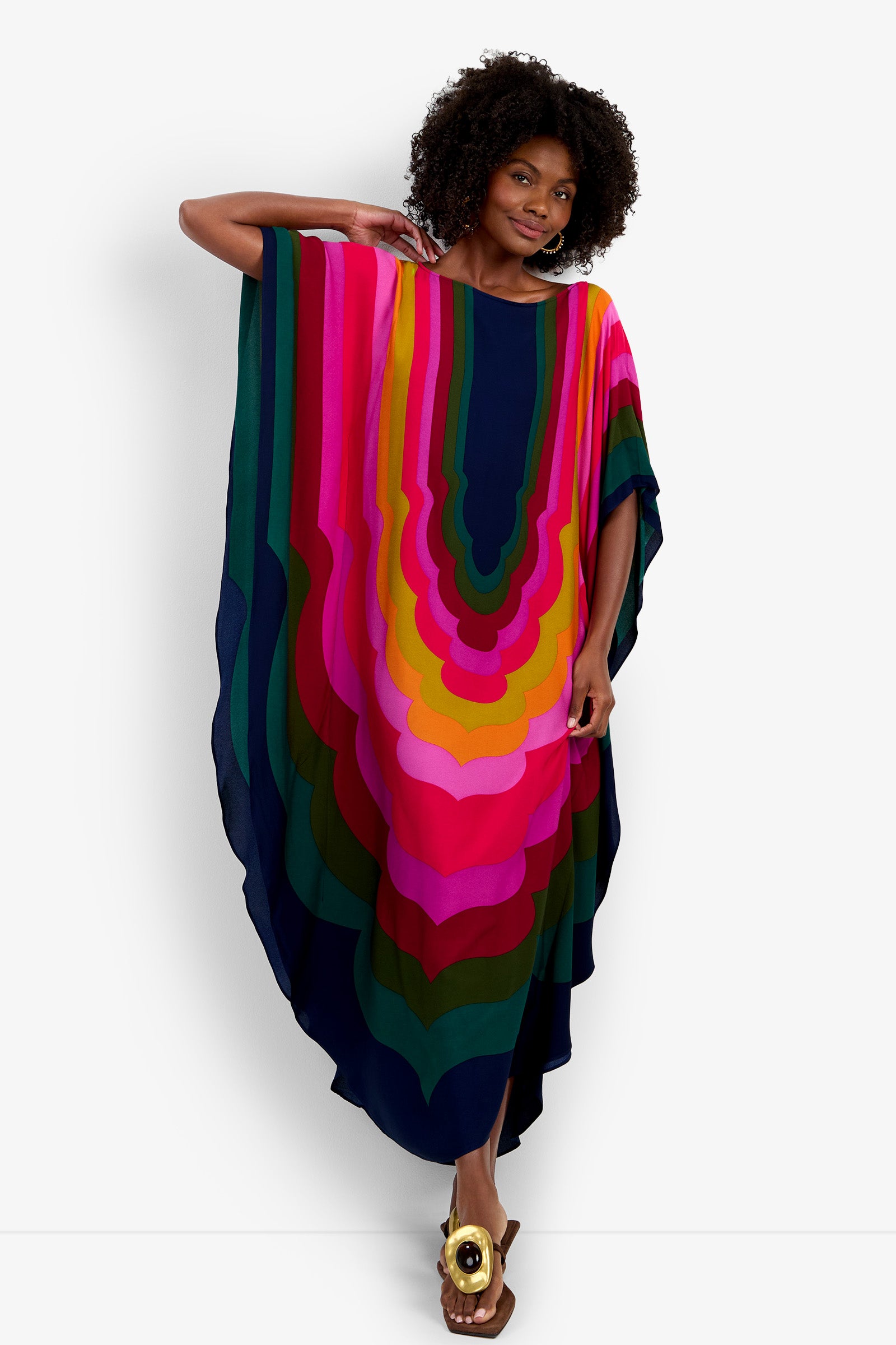 A woman with curly hair models the Vintage Moroccan Geo Caftan, barefoot and smiling. The bold, flowing caftan features wavy stripes in pink, orange, dark blue, green, and purple as she poses with one arm raised behind her head.