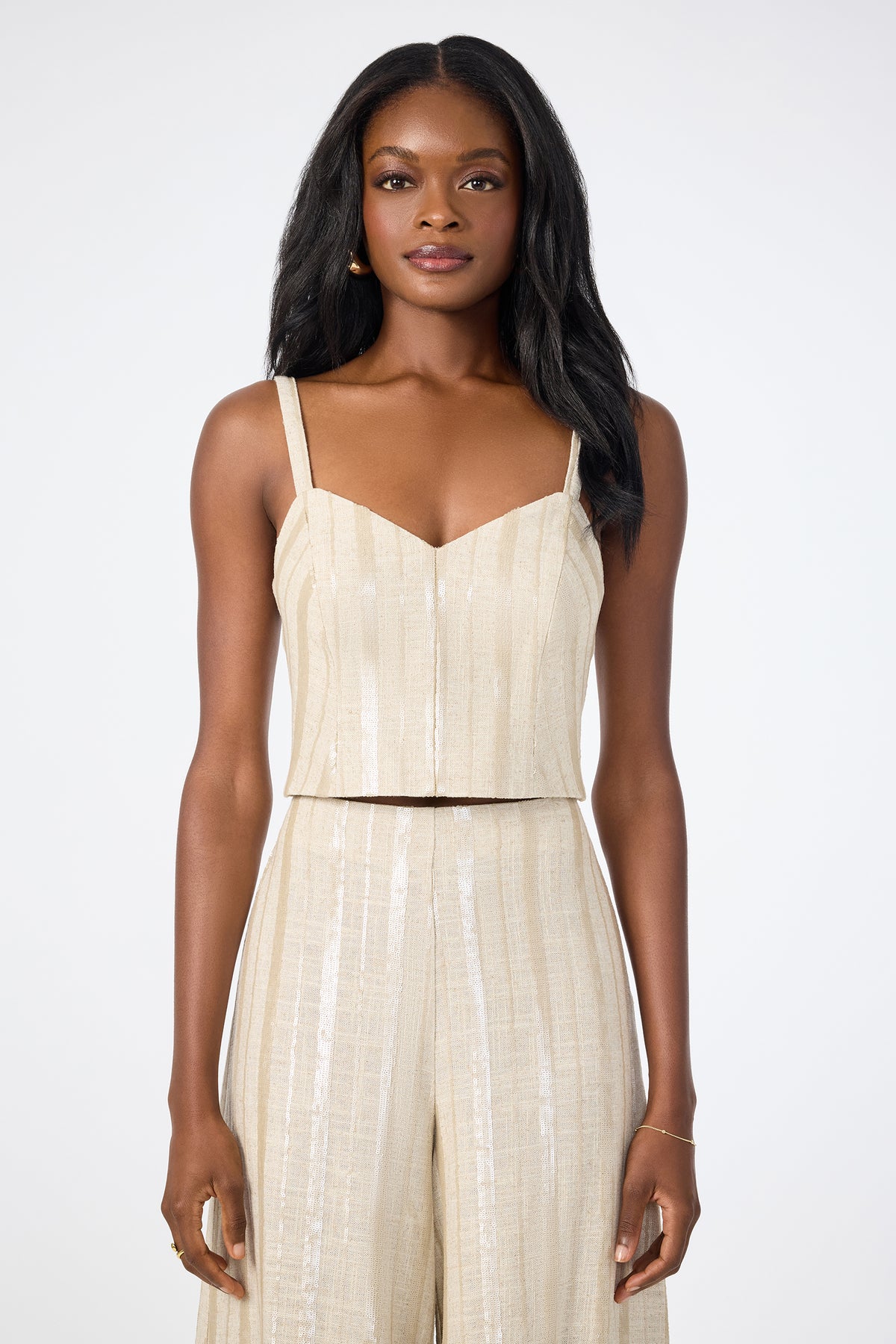 A woman with long, dark hair wears the Millau Sleeveless Top‚Äö√Ñ√Æbeige, textured, and cropped‚Äö√Ñ√Æpaired with matching high-waisted pants while standing against a plain white background.