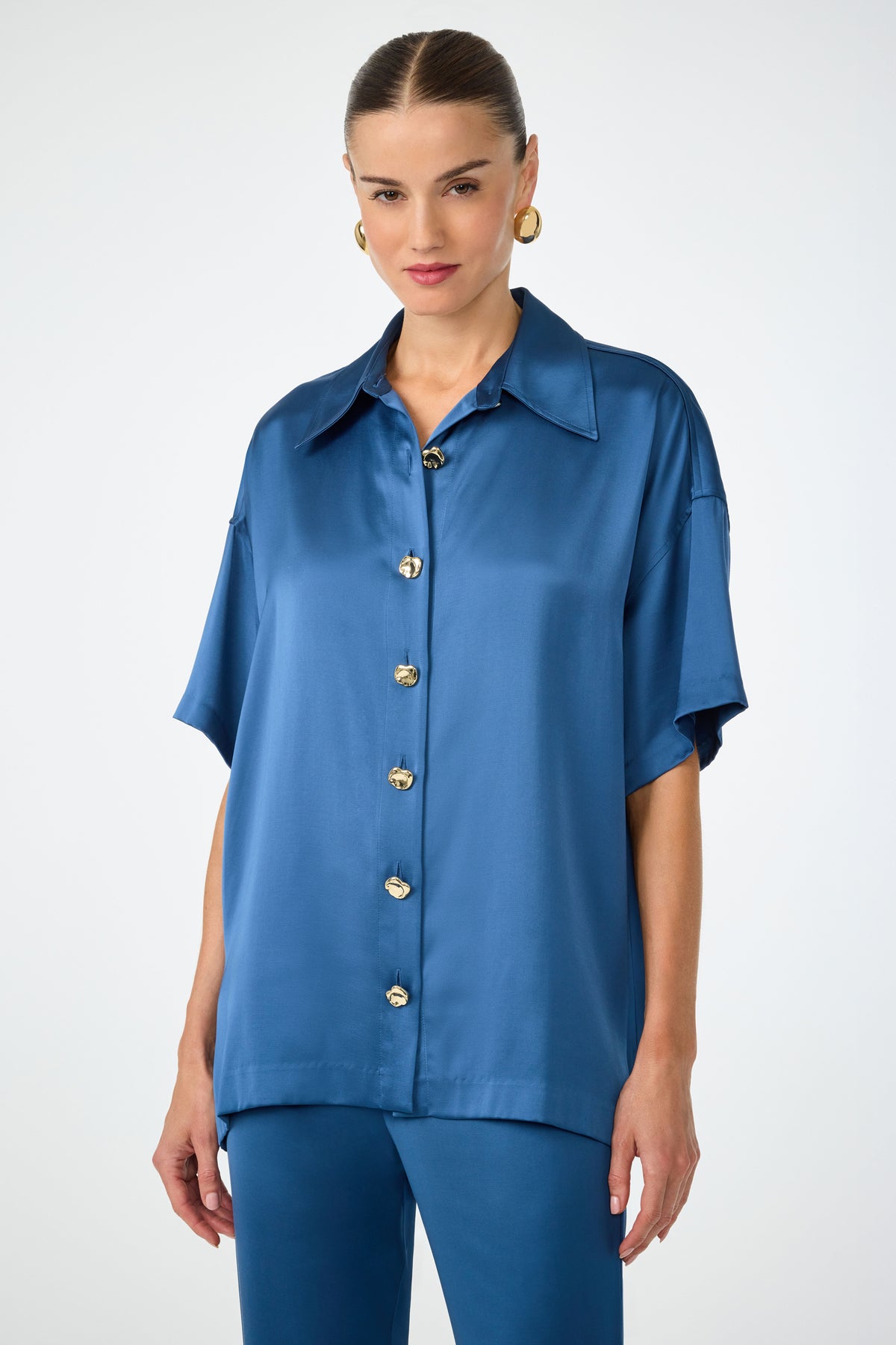 A woman wears the Manju Button-Front Top in Sumi Blue, styled with matching pants, gold button accents, large gold hoop earrings, and slicked-back hair against a plain light background.