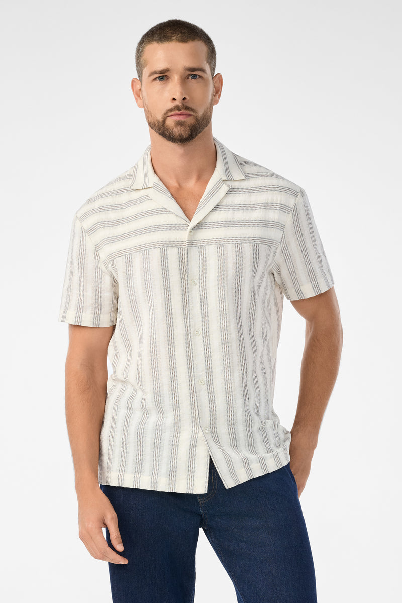 A man with short hair and a trimmed beard wears the Angelo Short-Sleeve Shirt - Stripe Gauze in Ecru, paired with dark blue jeans. He stands against a plain white background.