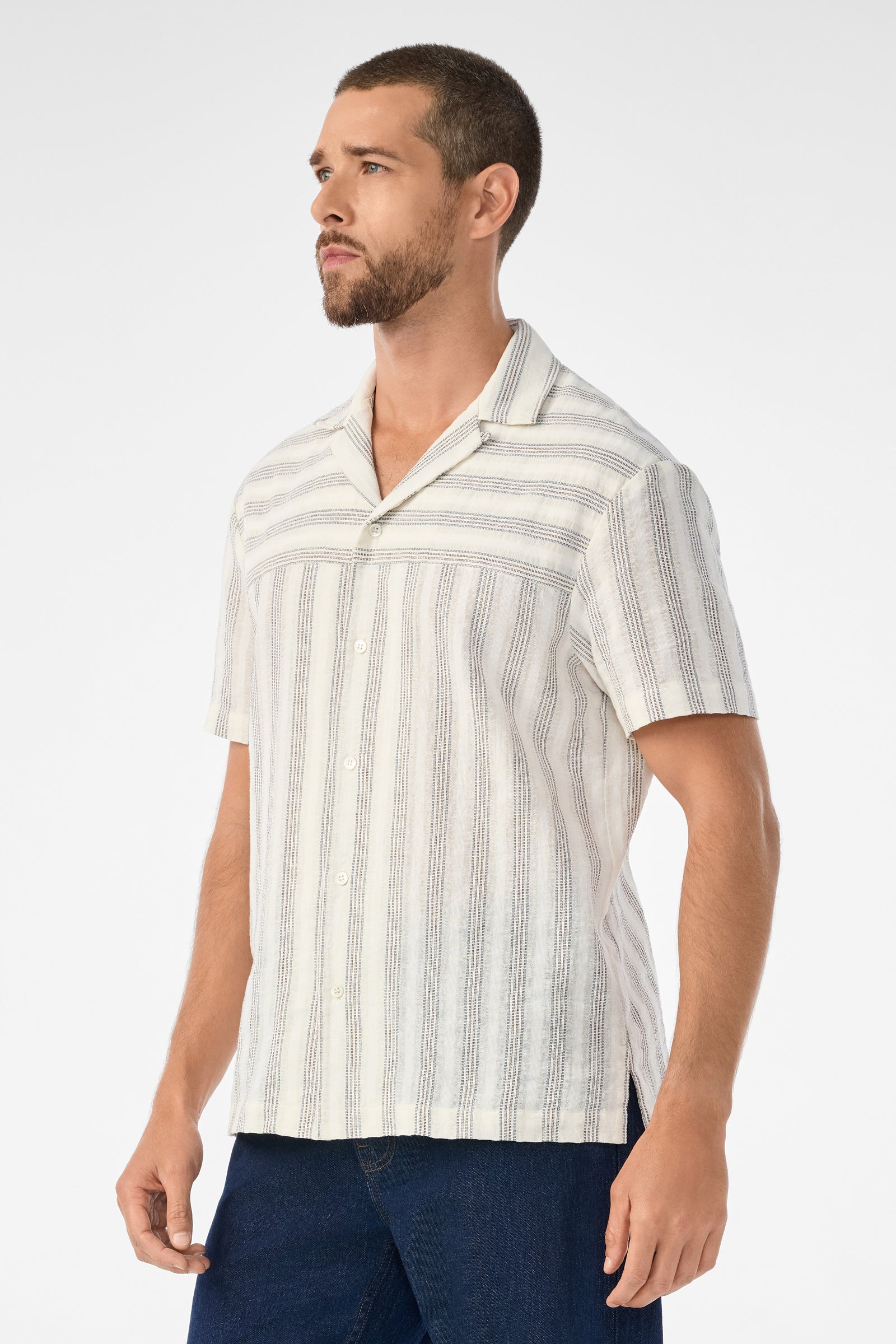 A man with short hair and a trimmed beard wears the Angelo Short-Sleeve Shirt - Stripe Gauze, a light-colored, vertically striped button-up, paired with dark blue pants, standing against a plain white background.