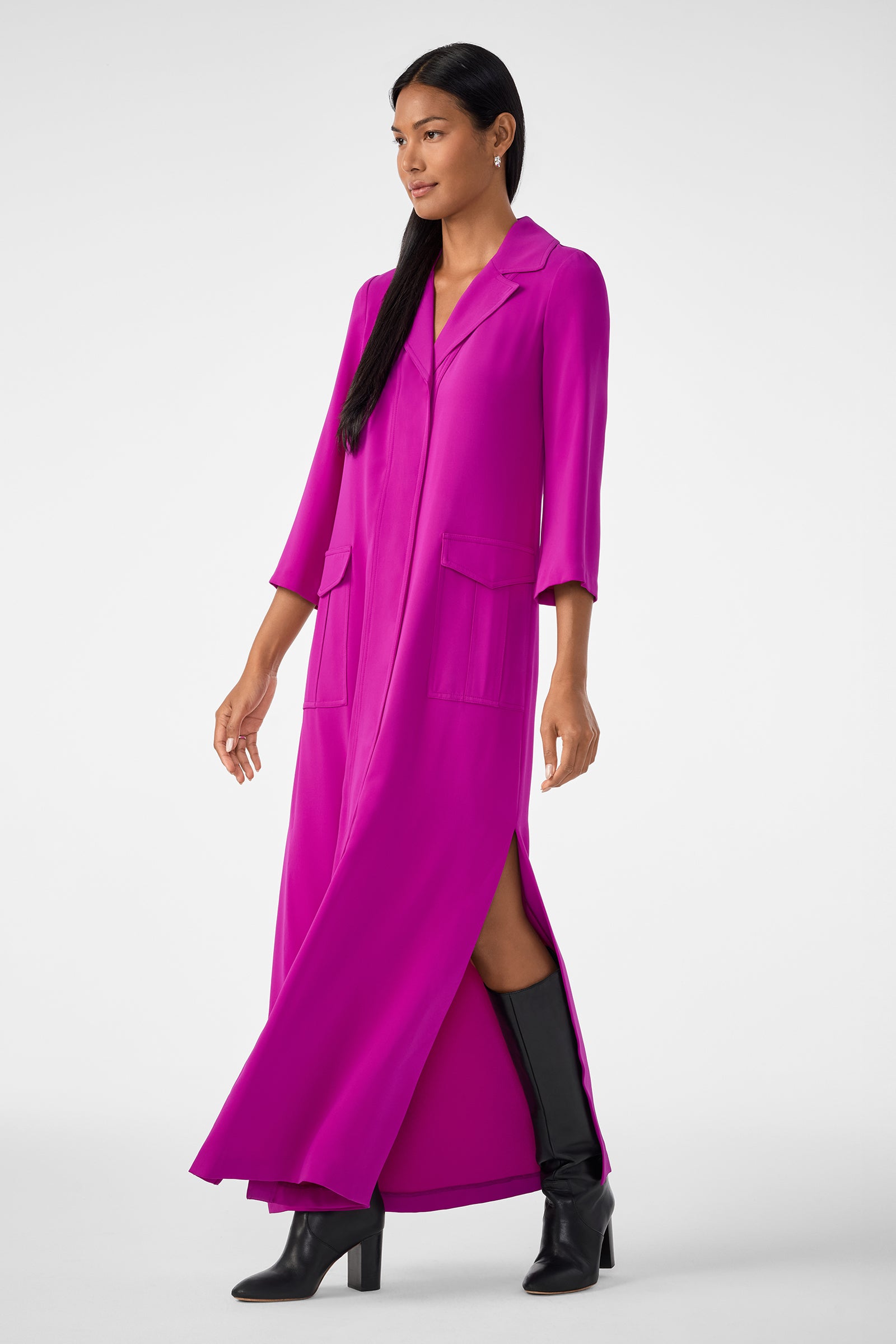 A woman wears the Annecy Dress—a long, bright magenta shirt dress with a collar, front pockets, three-quarter sleeves, and a side slit—paired with black knee-high boots against a plain light background.